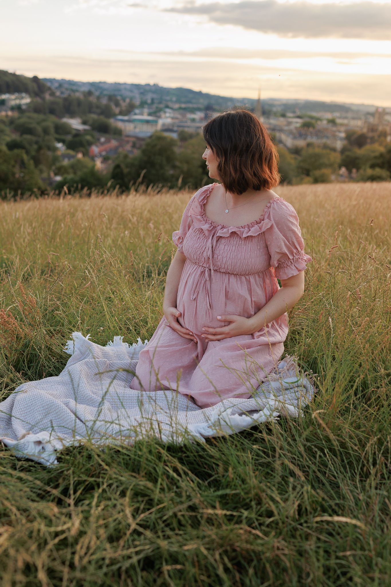 Maternity Photoshoot, woman in pink dress sits on blanket in grassy field with town view at dusk in Woodstock, Windsor.
