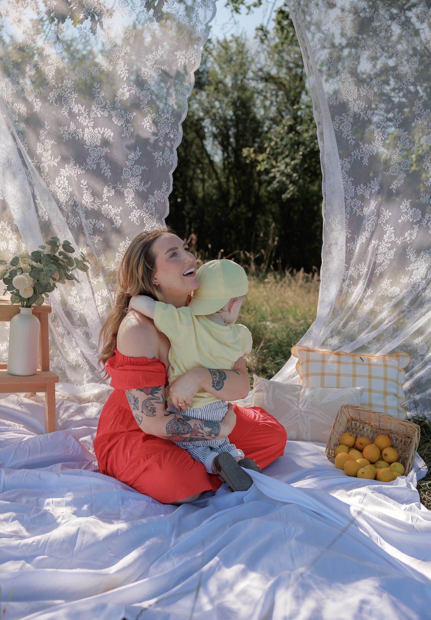 A smiling woman in red outfit holds a baby in yellow hat on a white blanket outdoors with lace curtains in Fairford, Cotswolds.