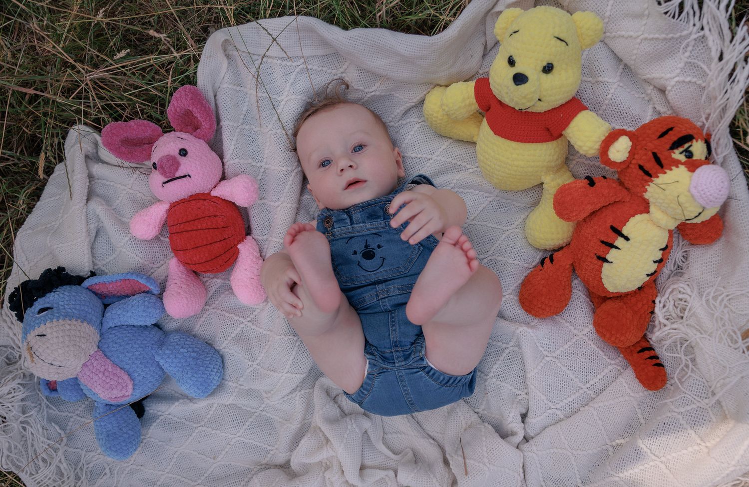 Baby in denim overalls lying on blanket surrounded by Winnie the Pooh stuffed animals including Piglet, Eeyore, Pooh, and Tigger in Burford.