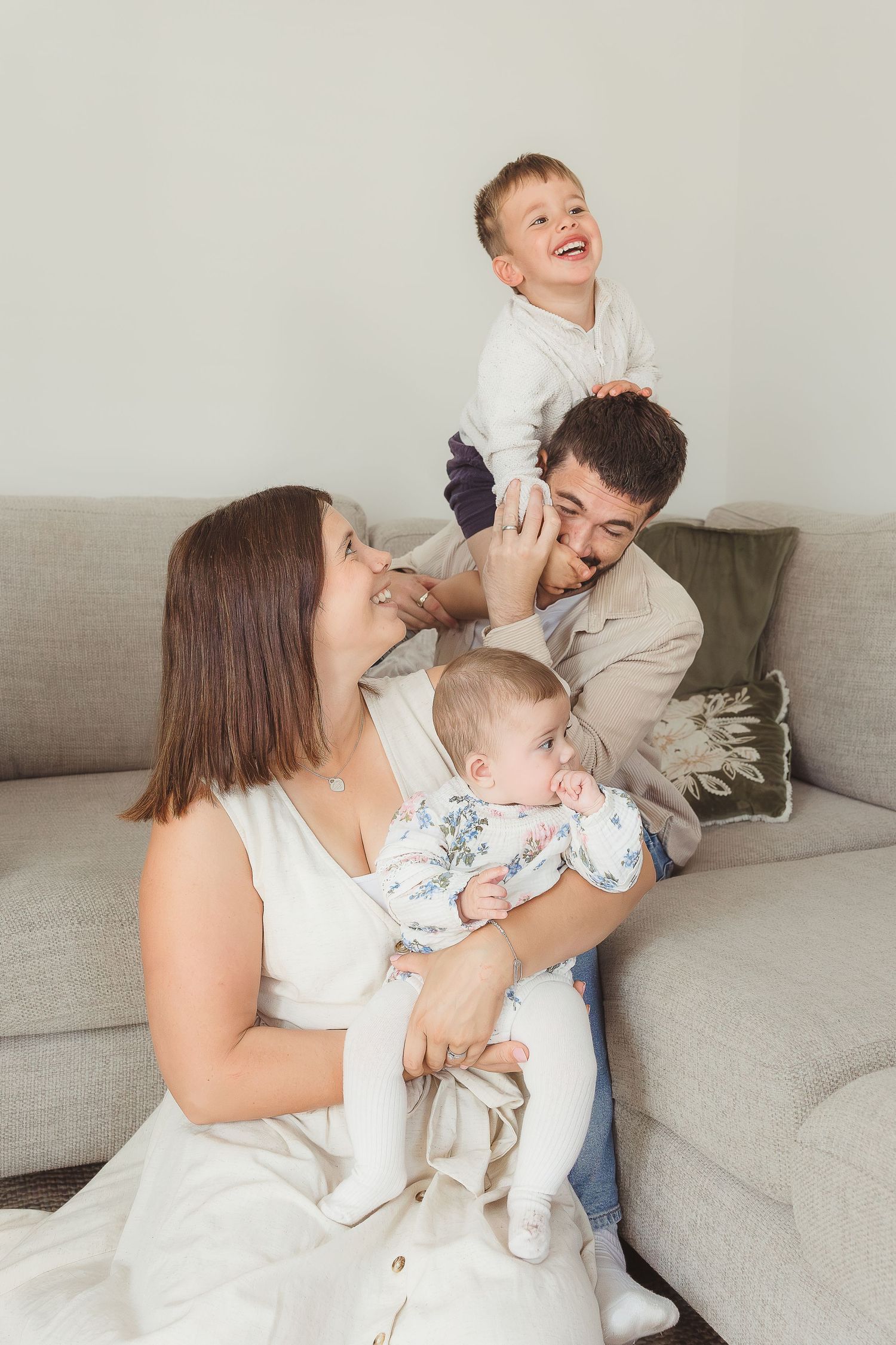 Happy family with two children relaxing on a cozy gray sofa, parents playing with toddler and holding newborn baby  in Broadway, Cotswolds.