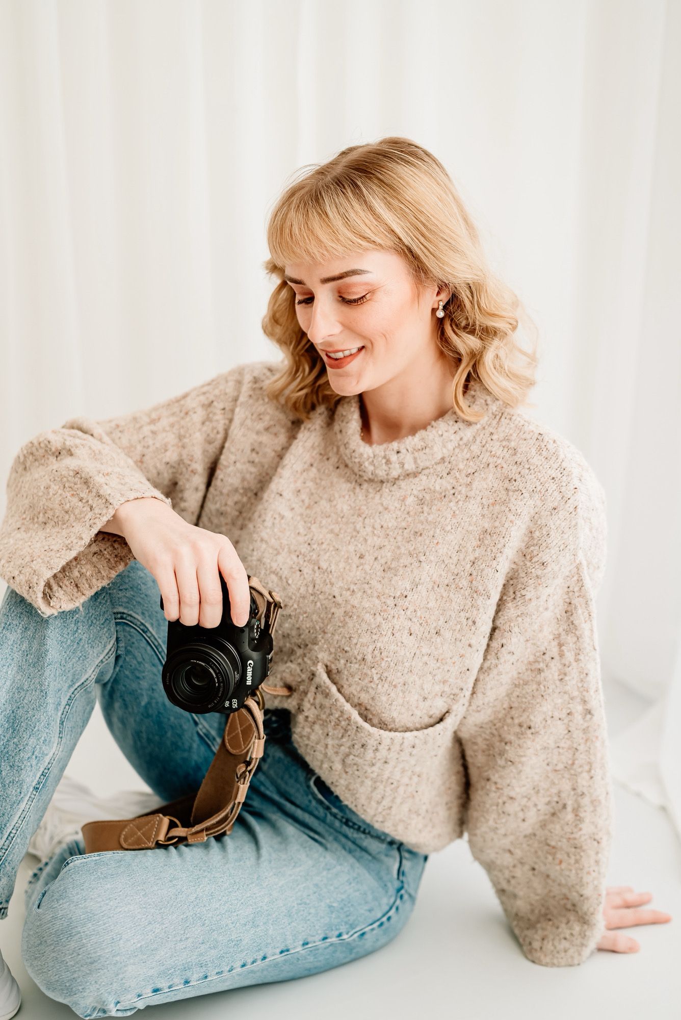 Person in beige sweater and jeans holds camera while posing against white background in Bath, Somerset.