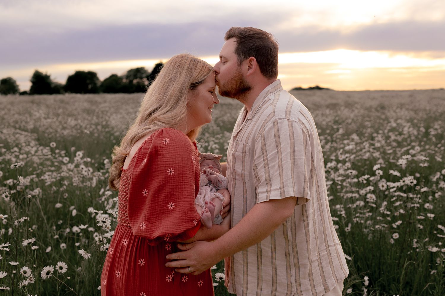 Couple sharing a romantic kiss at sunset in a blooming daisy field, woman in red dress holding flowers.