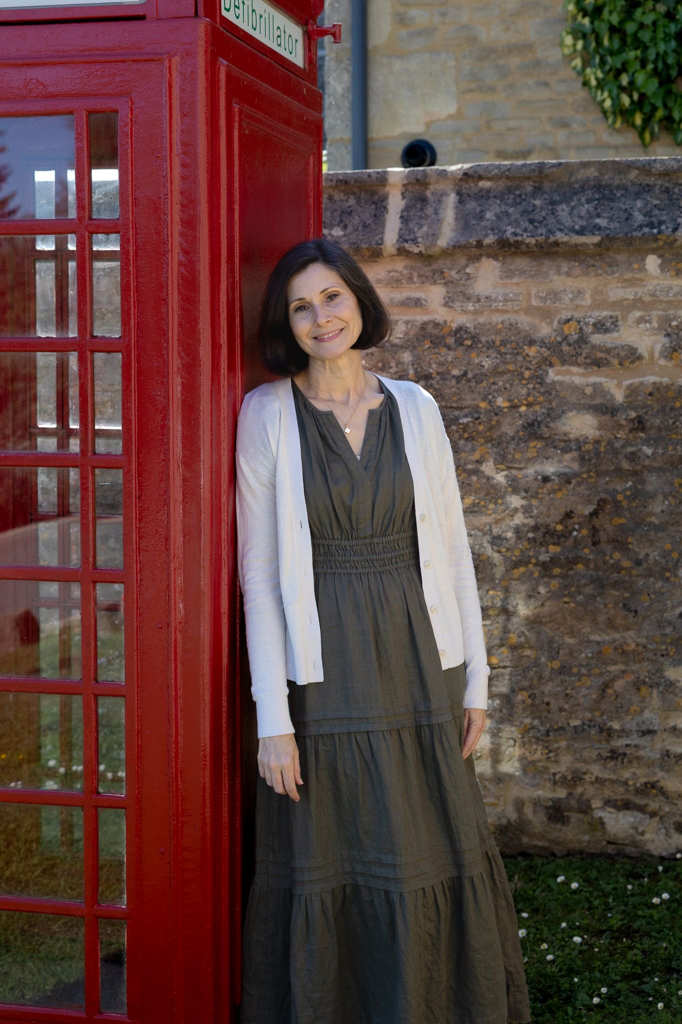 Woman in olive dress and white cardigan smiling beside a classic red British telephone box.
