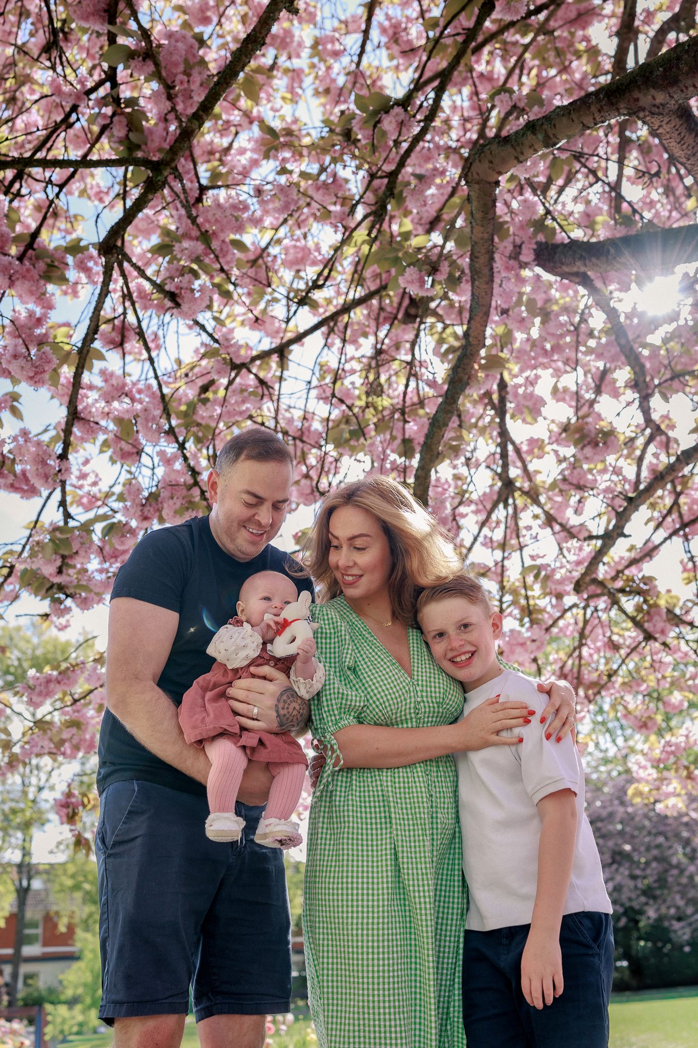 Family of four posing under pink cherry blossom tree, parents holding baby and hugging young boy in spring garden in Bristol.