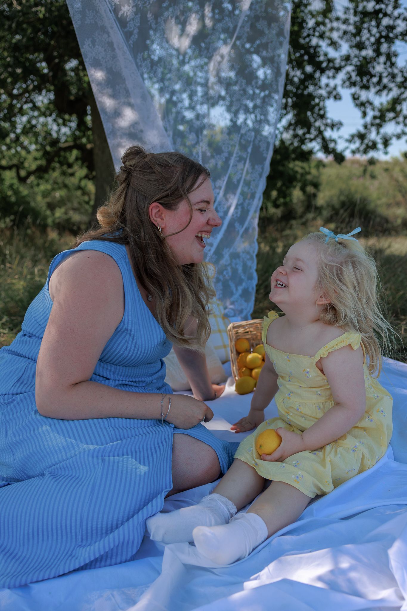 Woman in blue dress and toddler girl in yellow dress laughing together at a sunny outdoor picnic in South Cerney, Cotswolds.