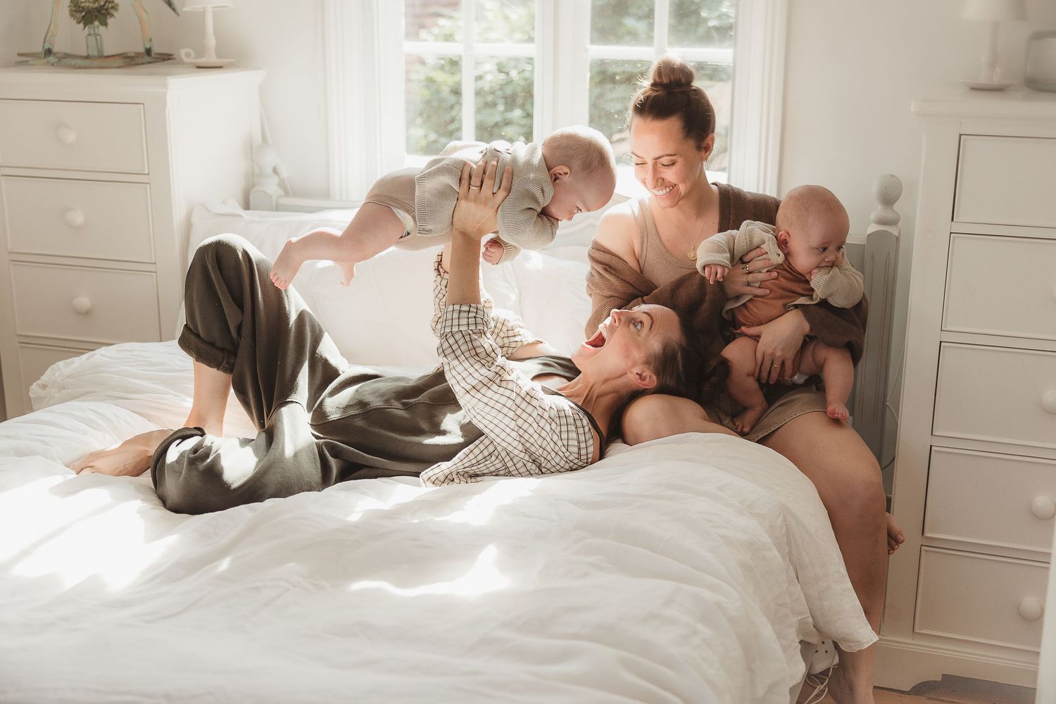 Same sex couple playfully interact with their babies on a bright white bed in a sunlit bedroom in Great Tew, Cotswolds.