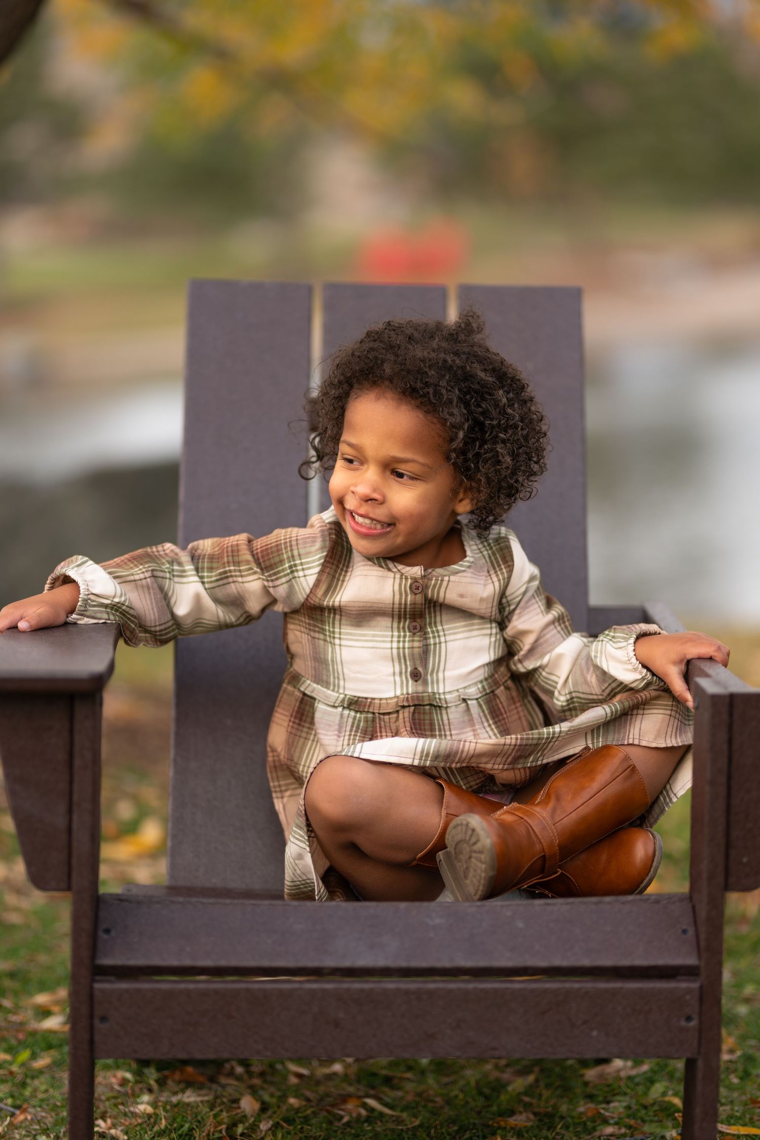 A young mixed-race girl in a plaid dress sits cross-legged in a large wooden chair outdoors in autumn.