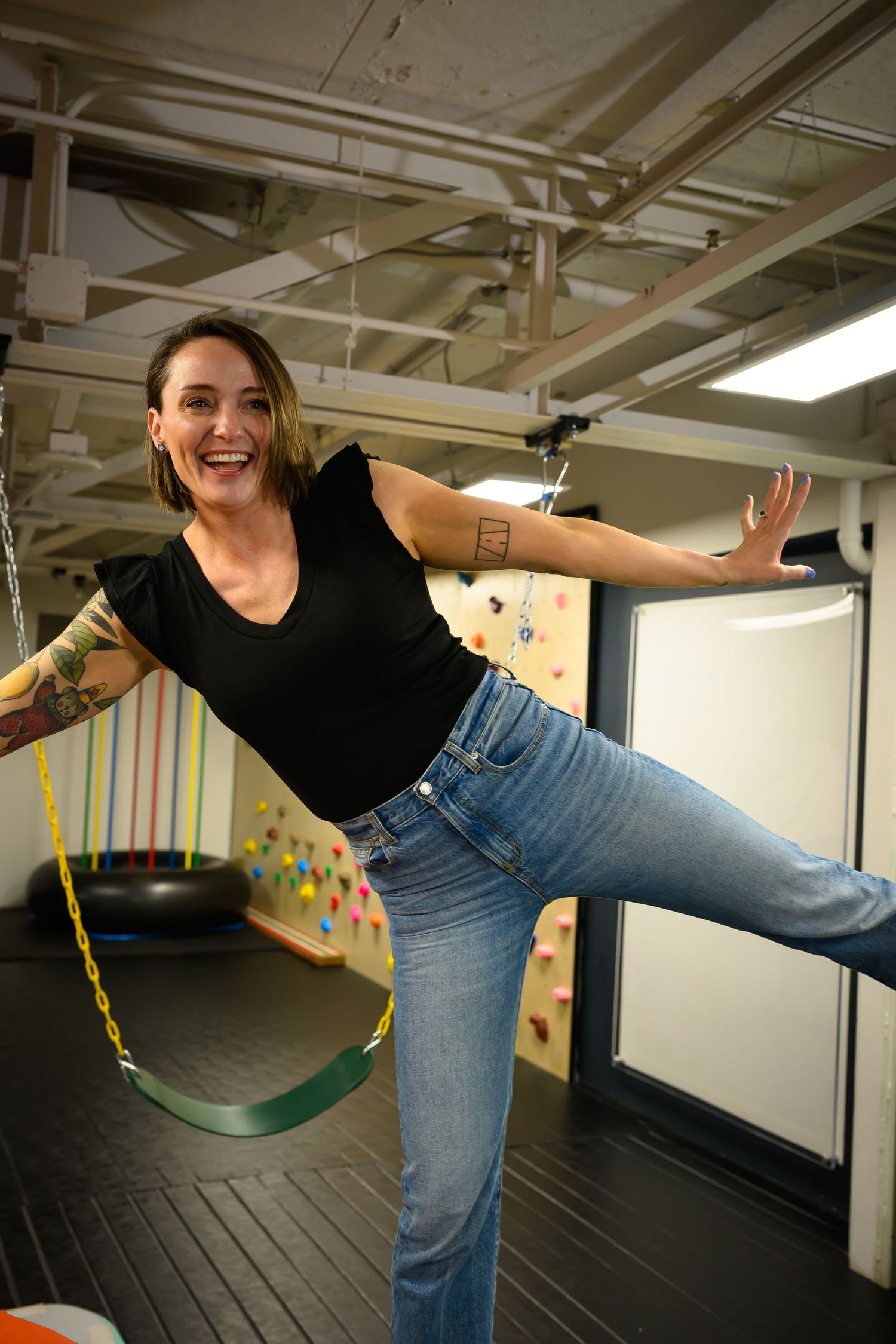 Smiling woman balancing on one leg near swing in colorful pediatric therapy gym.