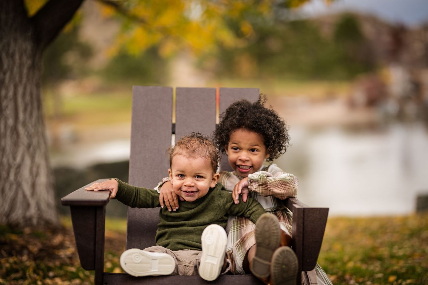 Two young mixed-race siblings sit together smiling in a wooden outdoor chair with autumn foliage behind them.