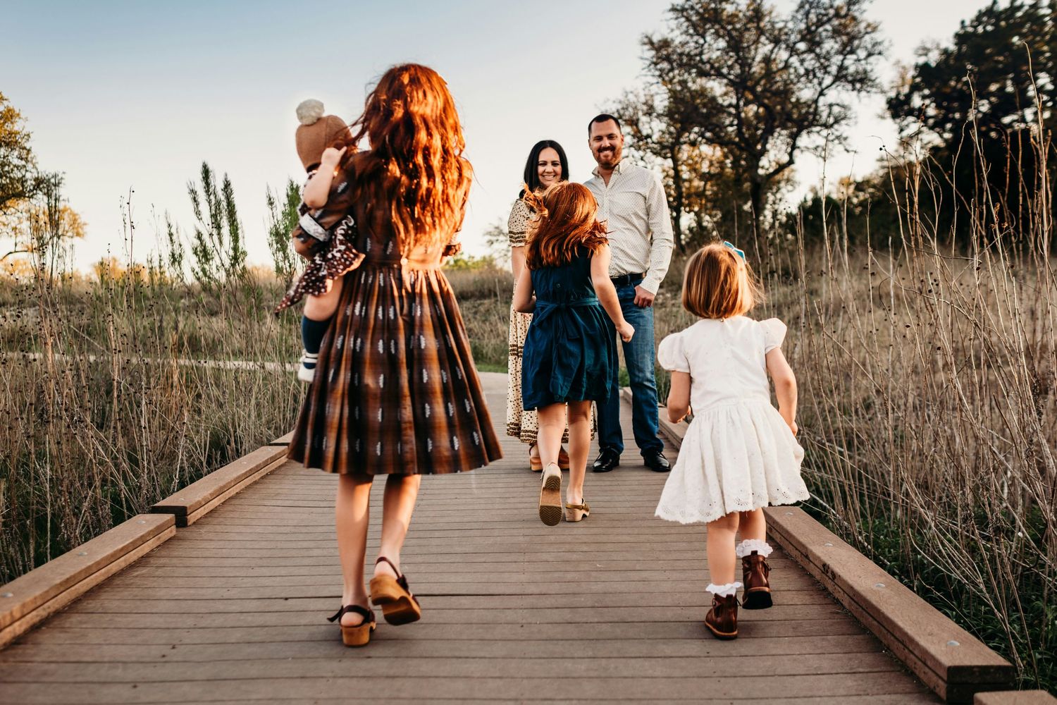 Family walking on a wooden boardwalk path through golden fields at sunset, children in dresses, parents smiling warmly.