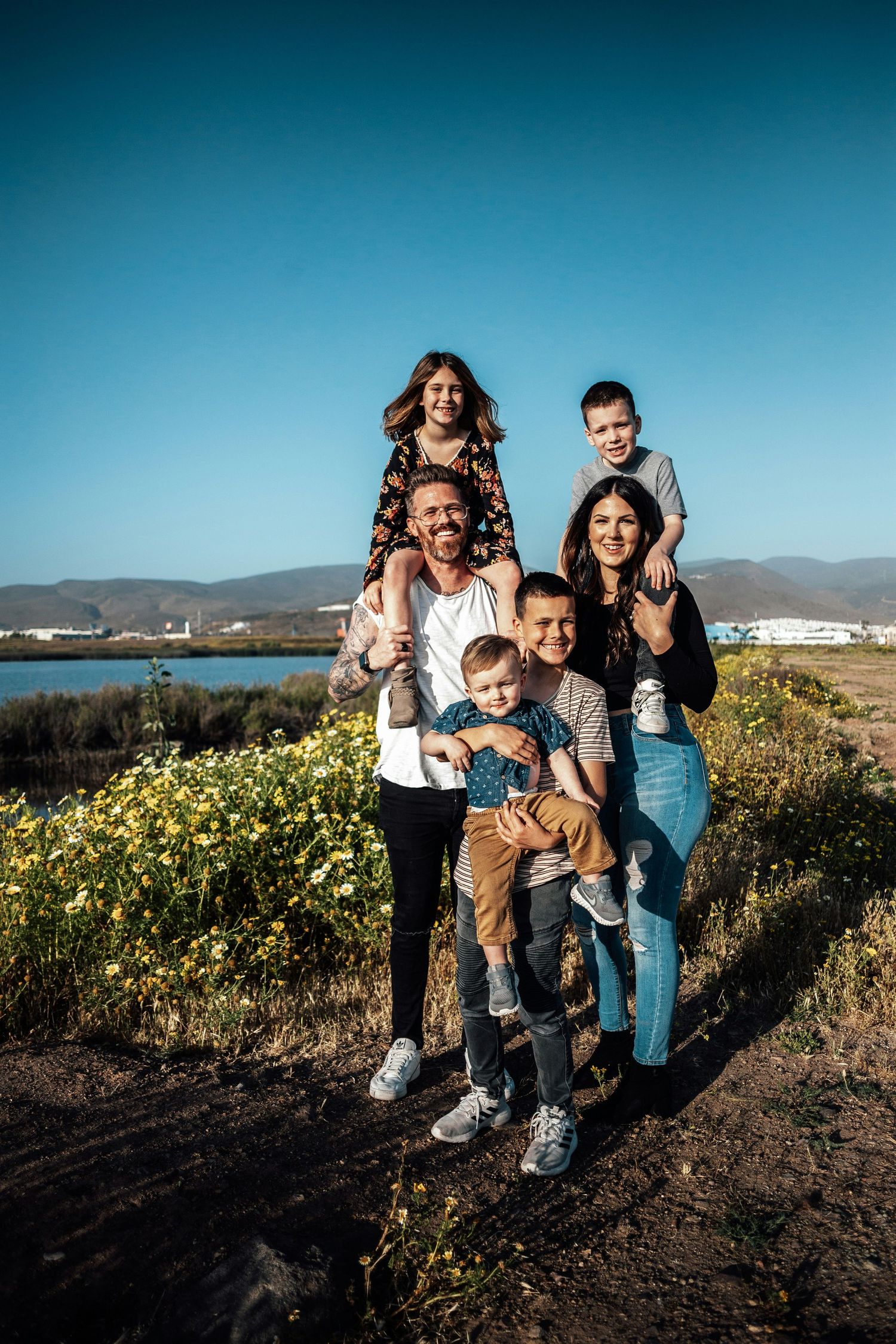 Happy family of six posing outdoors near a lake with yellow wildflowers and blue sky in background.