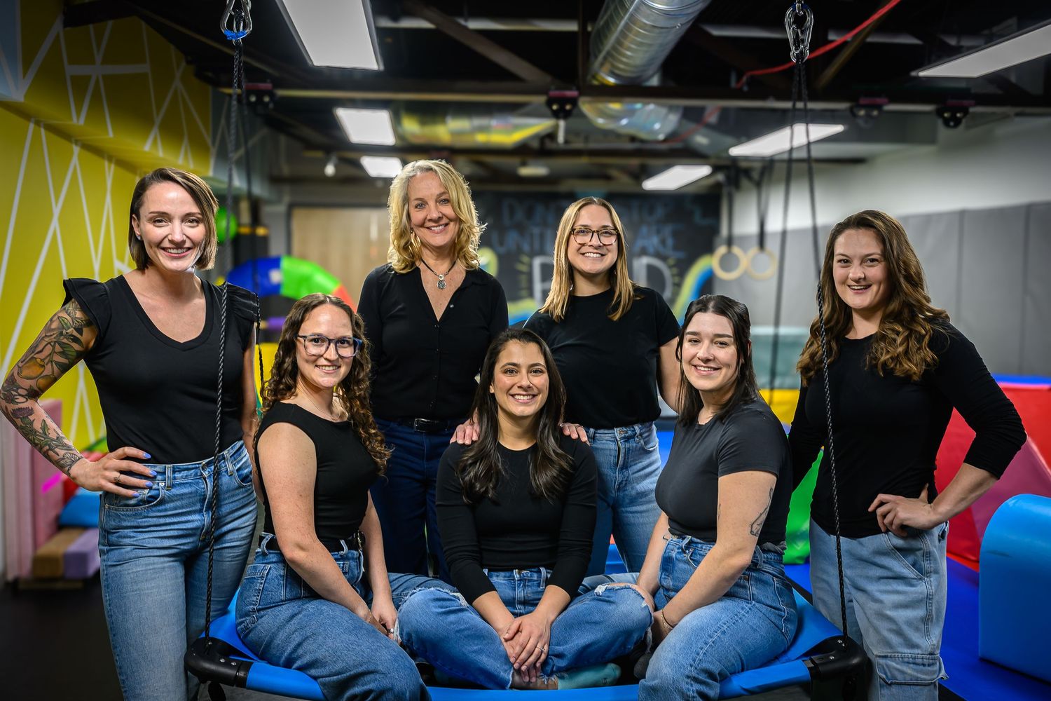 Seven women in matching black tops and jeans posing together in colorful indoor play facility.