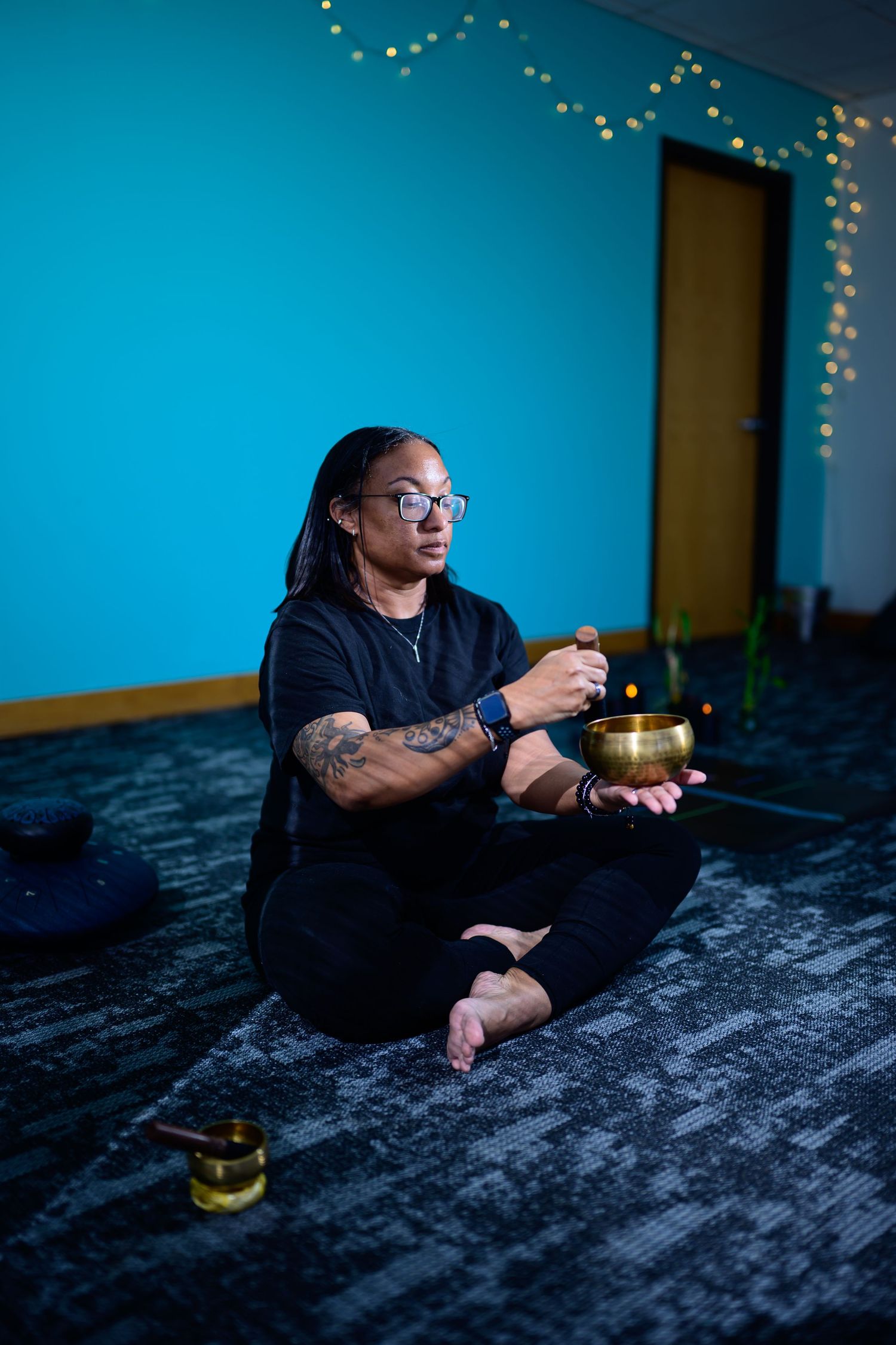 A tattooed man with dreadlocks and glasses sits cross-legged playing a Tibetan singing bowl in a blue meditation room.