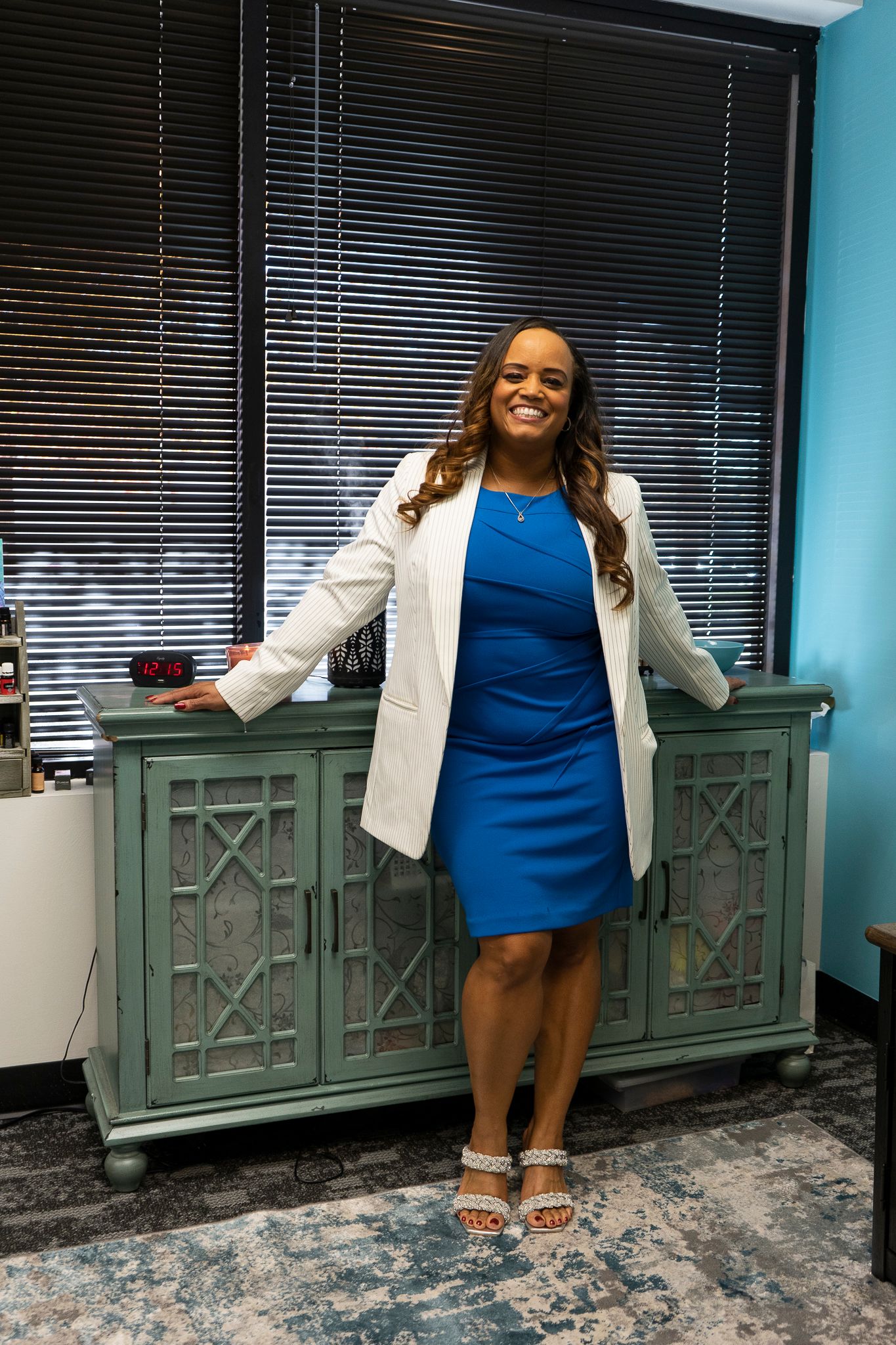 A smiling woman in a blue dress and white blazer poses confidently beside a teal cabinet in a professional office setting.