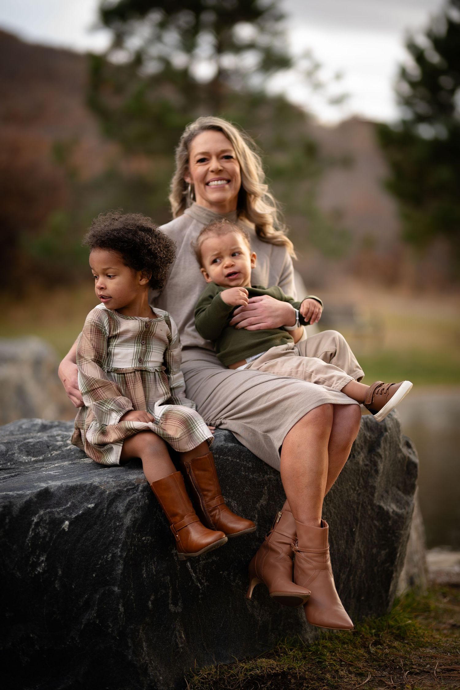 A smiling blonde mother sits on a large rock outdoors holding two young children in autumn clothing.