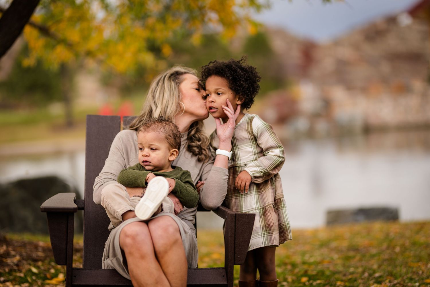 A blonde mother kisses her young daughter while holding a baby in a wooden chair by an autumn lake.