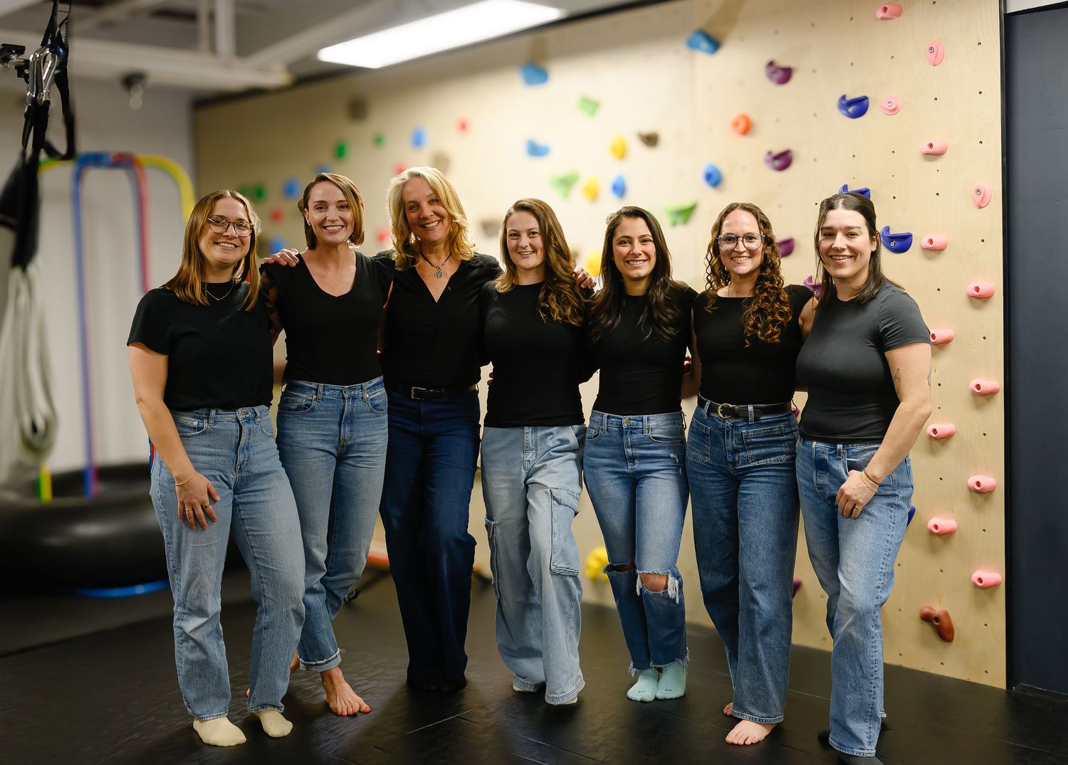 Seven women in matching black tops and jeans smiling together in front of a colorful climbing wall.