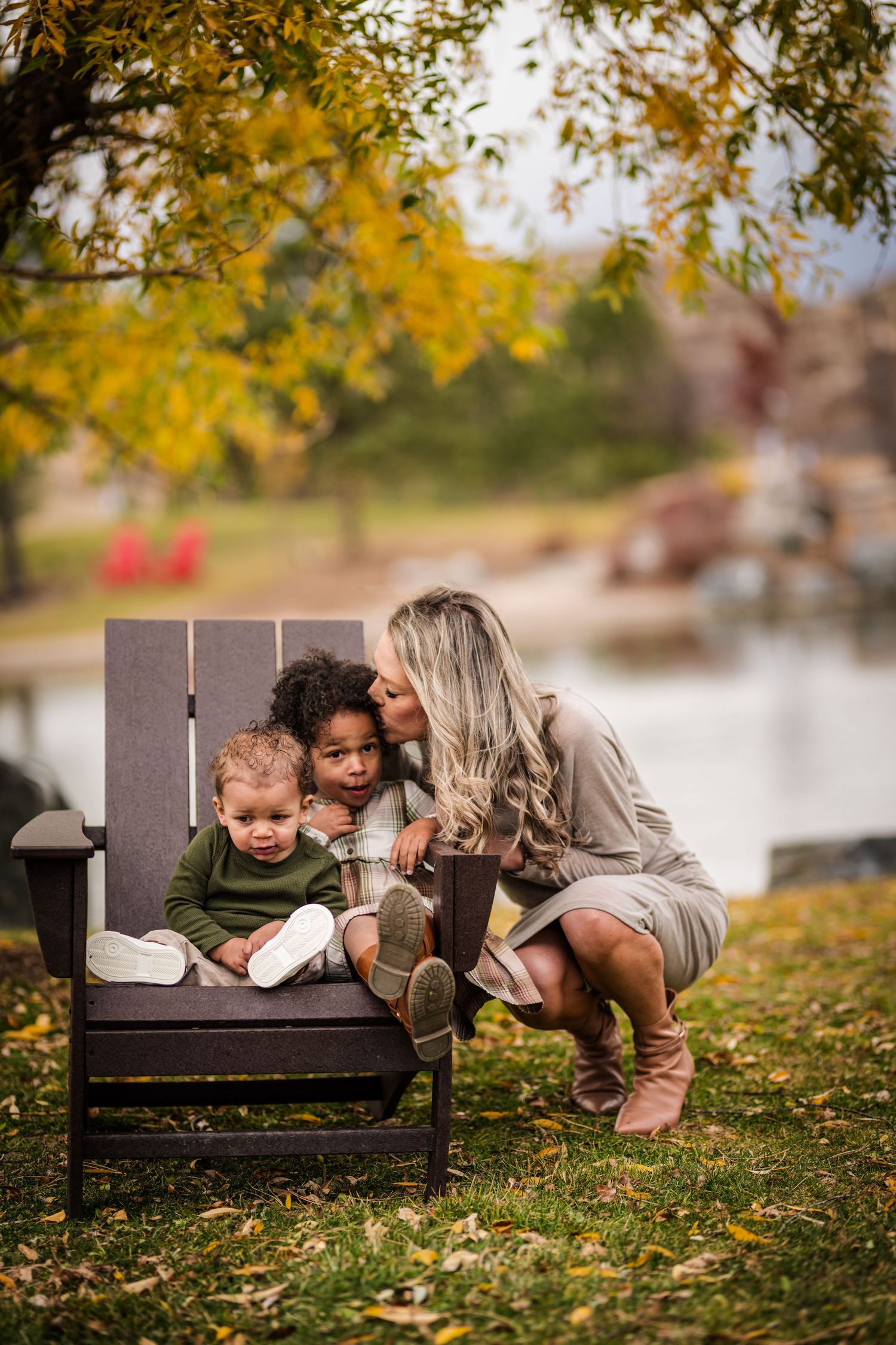 A blonde woman crouches beside a dark wooden chair holding two young children in a fall outdoor setting.