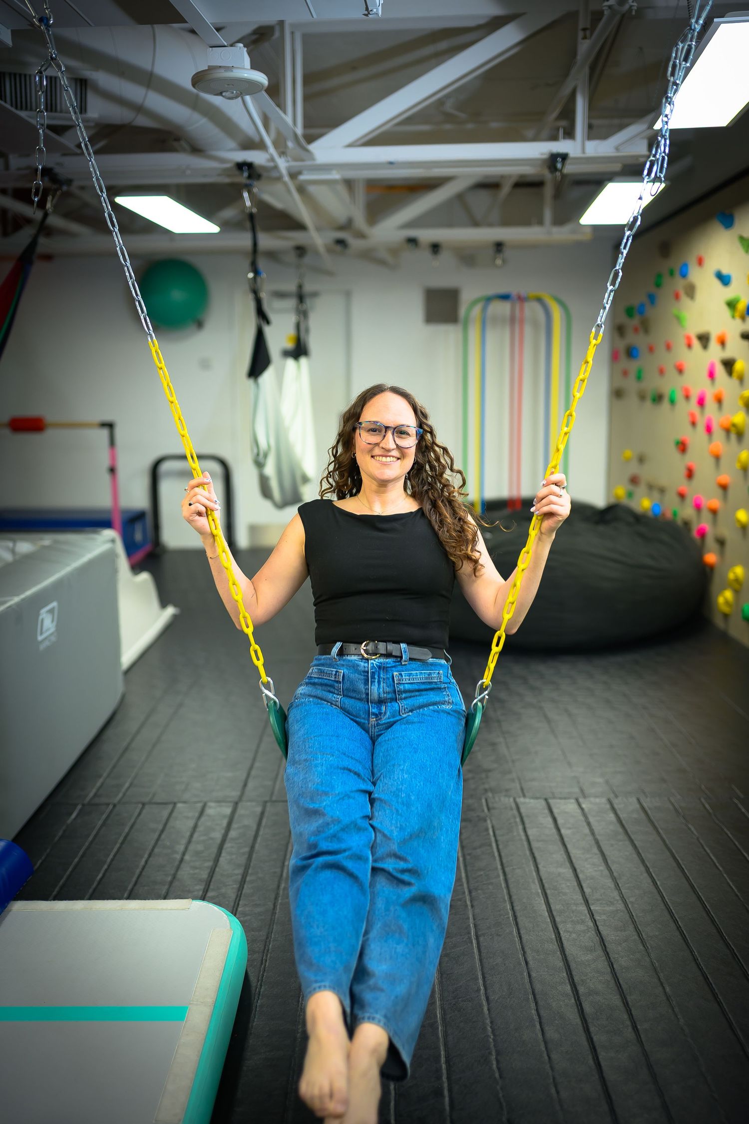 Woman smiling on yellow chain swing in colorful pediatric therapy gym with climbing wall.
