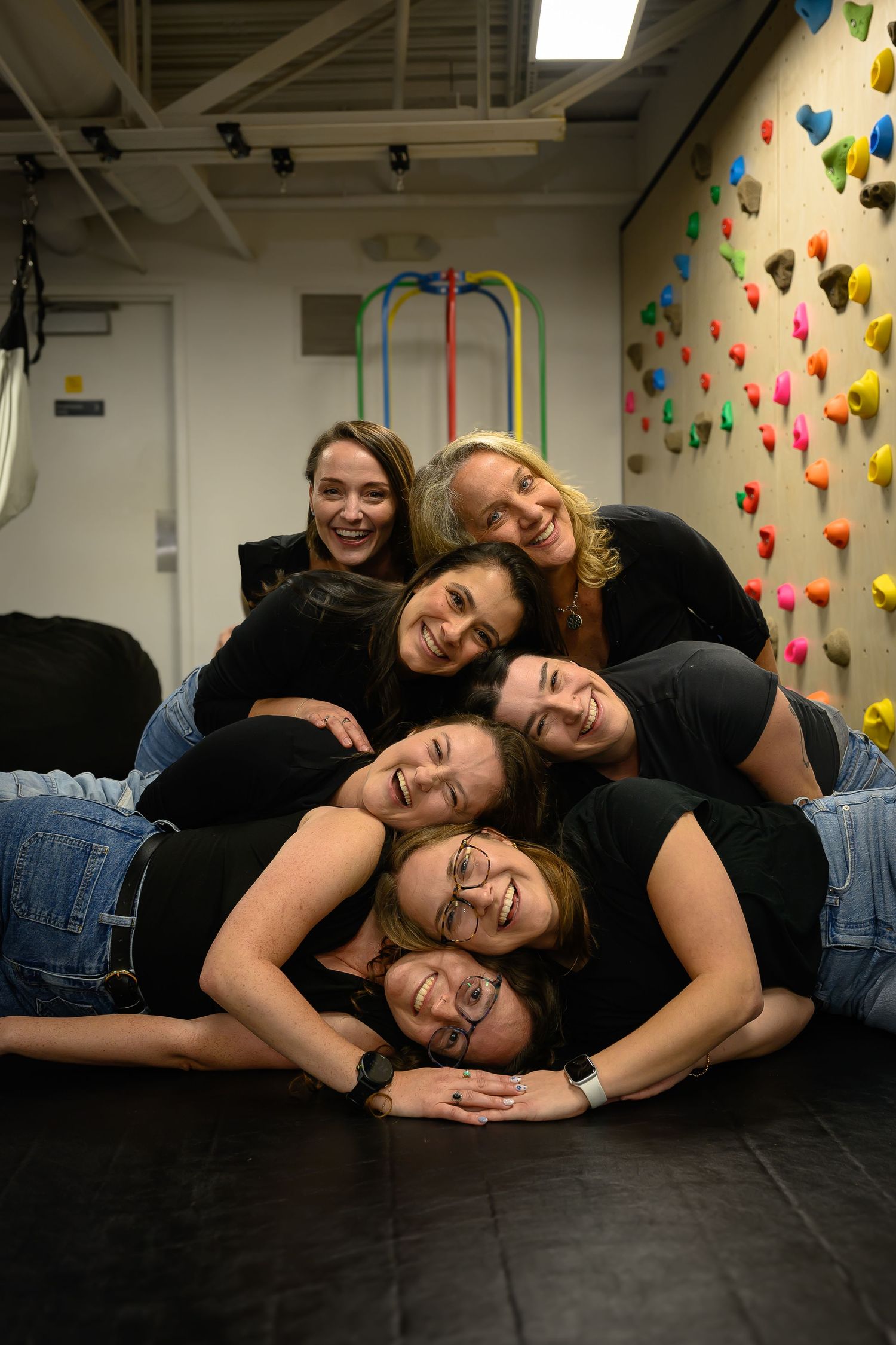 Group of six women in black tops and jeans playfully stacked together near colorful climbing wall.