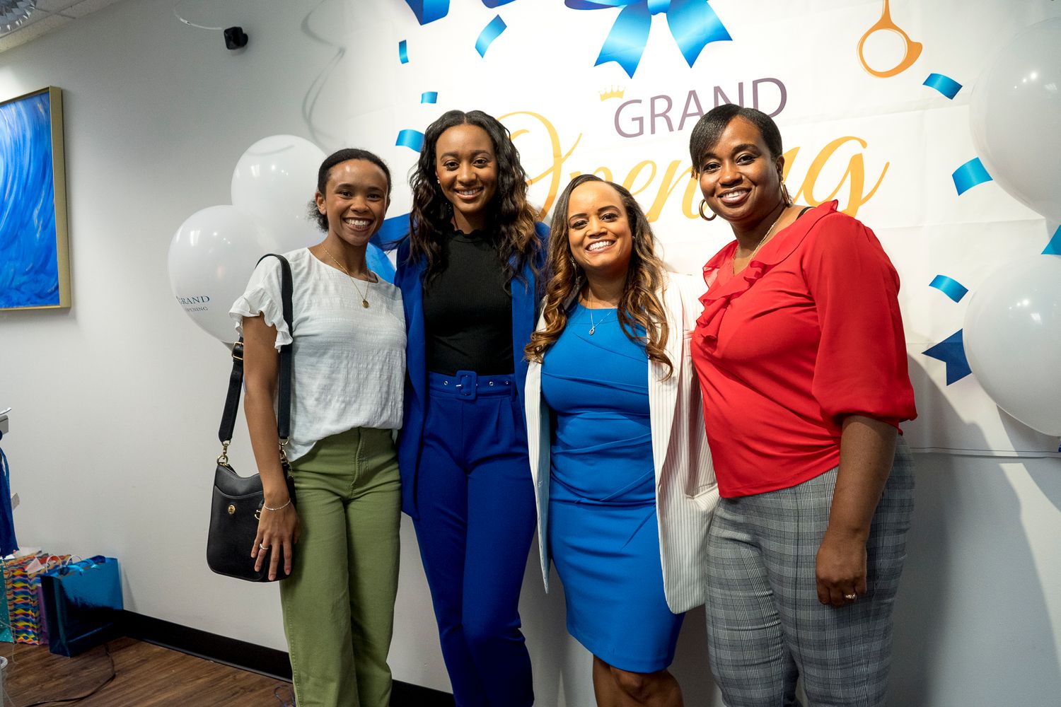 Four women smile together at grand opening celebration in front of festive balloon and ribbon backdrop.