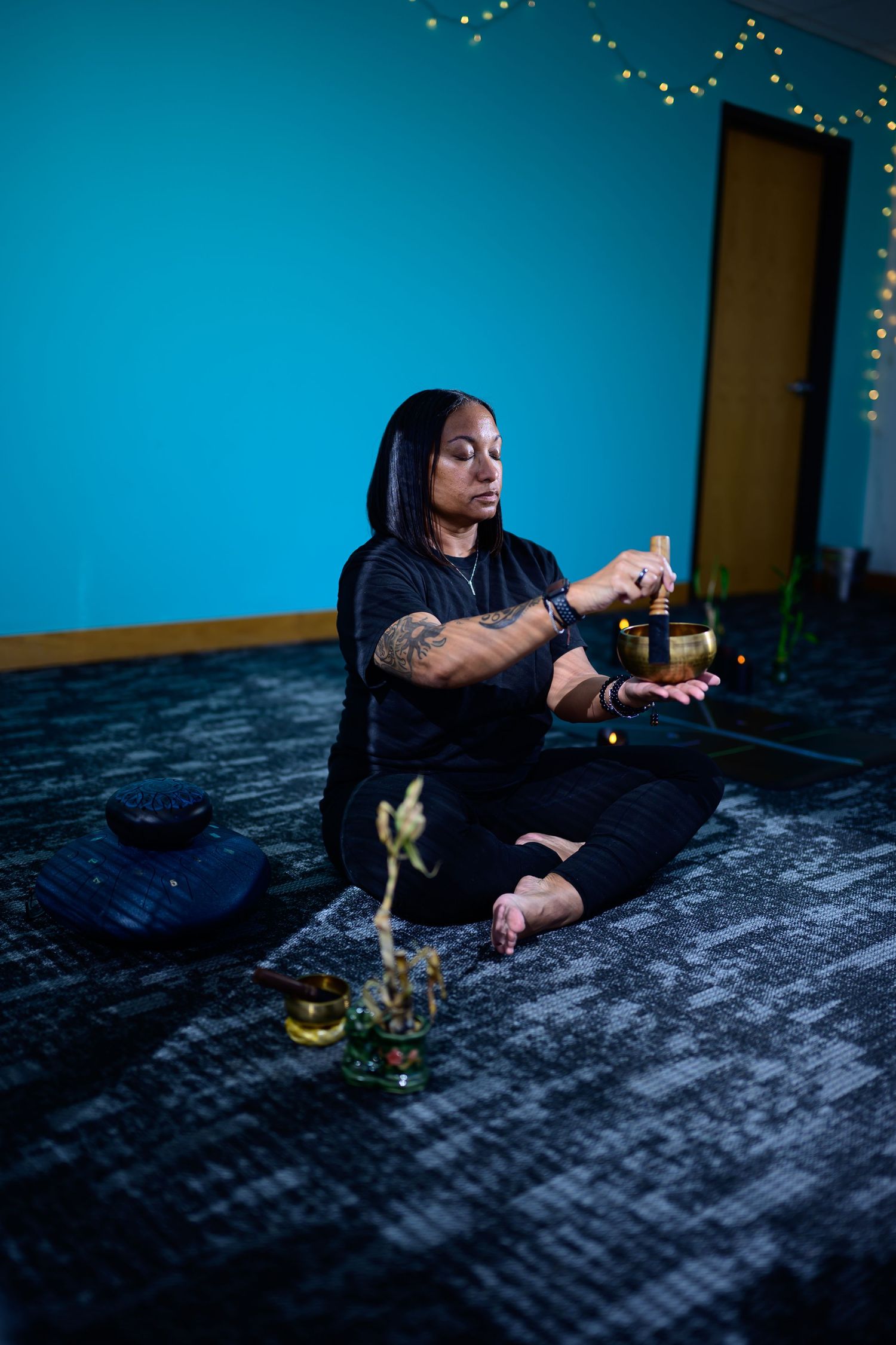 Woman in black outfit playing a Tibetan singing bowl while seated cross-legged on floor in blue meditation room.
