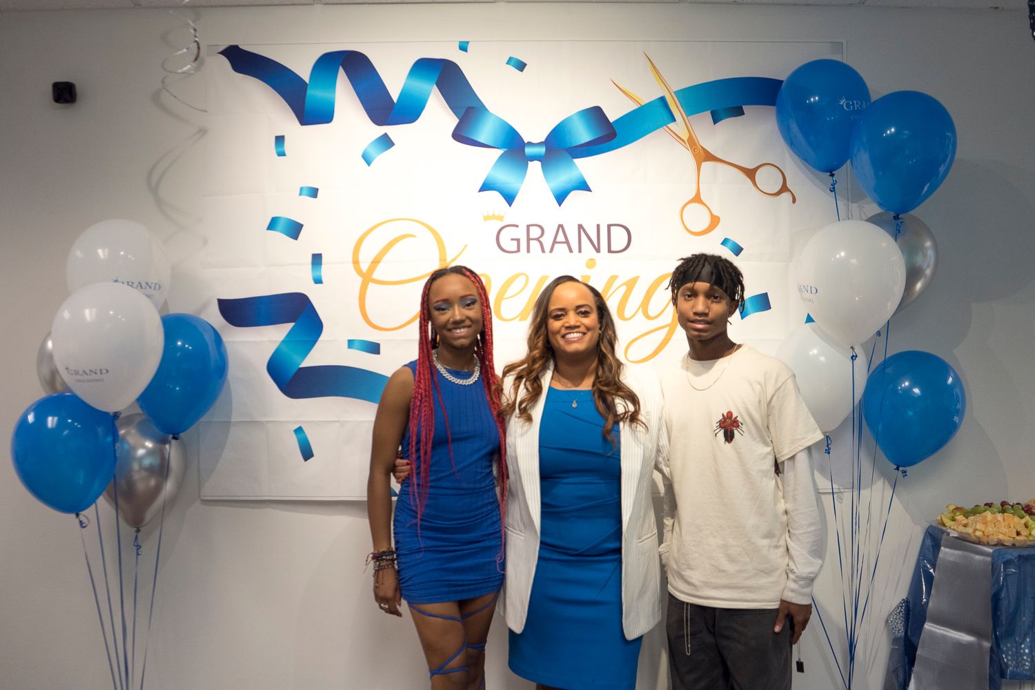 Three people celebrate grand opening event posing in front of decorative ribbon and balloon display.