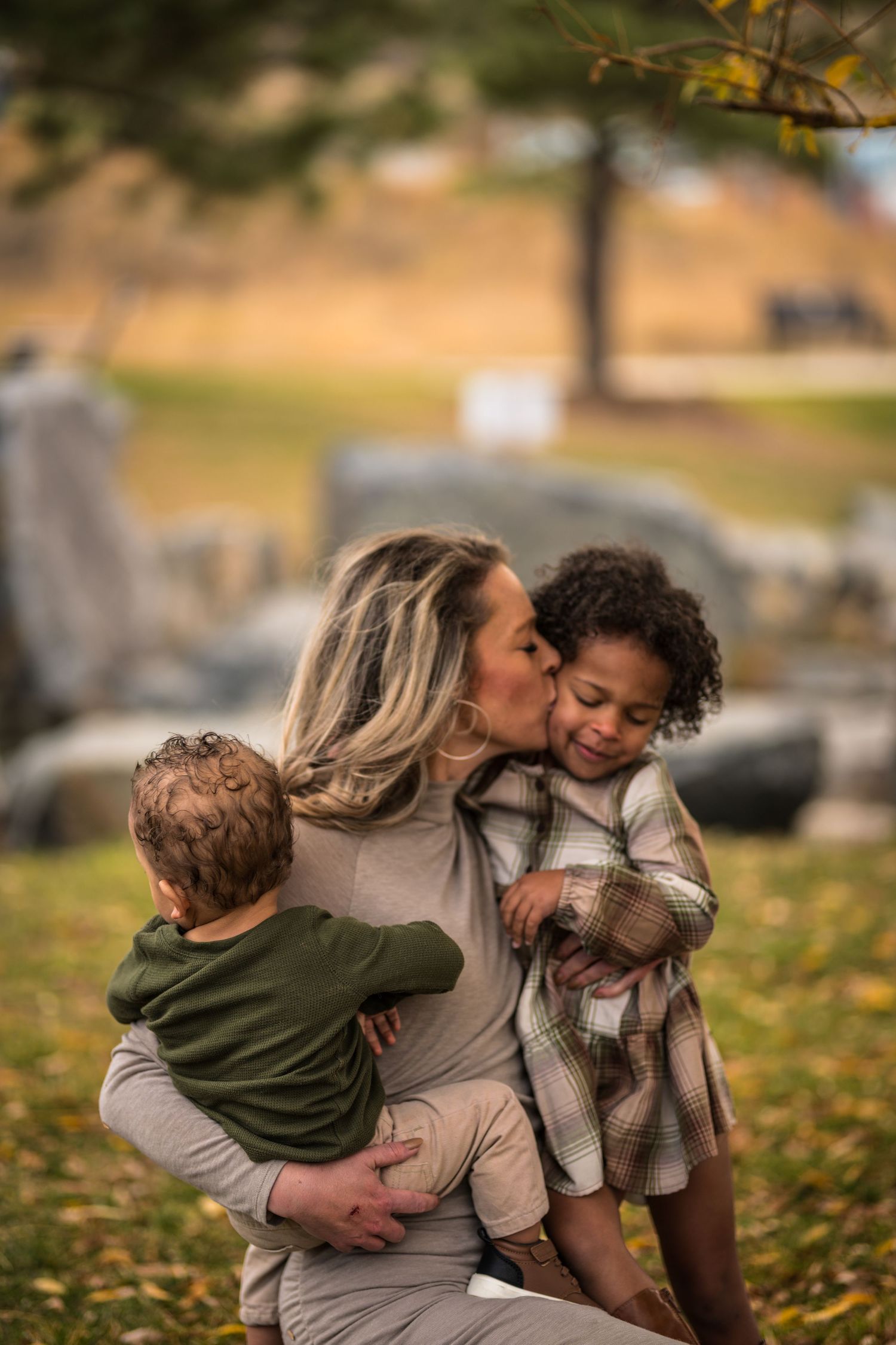 A mother kisses her young daughter on the cheek while holding two children outdoors in an autumn park setting.