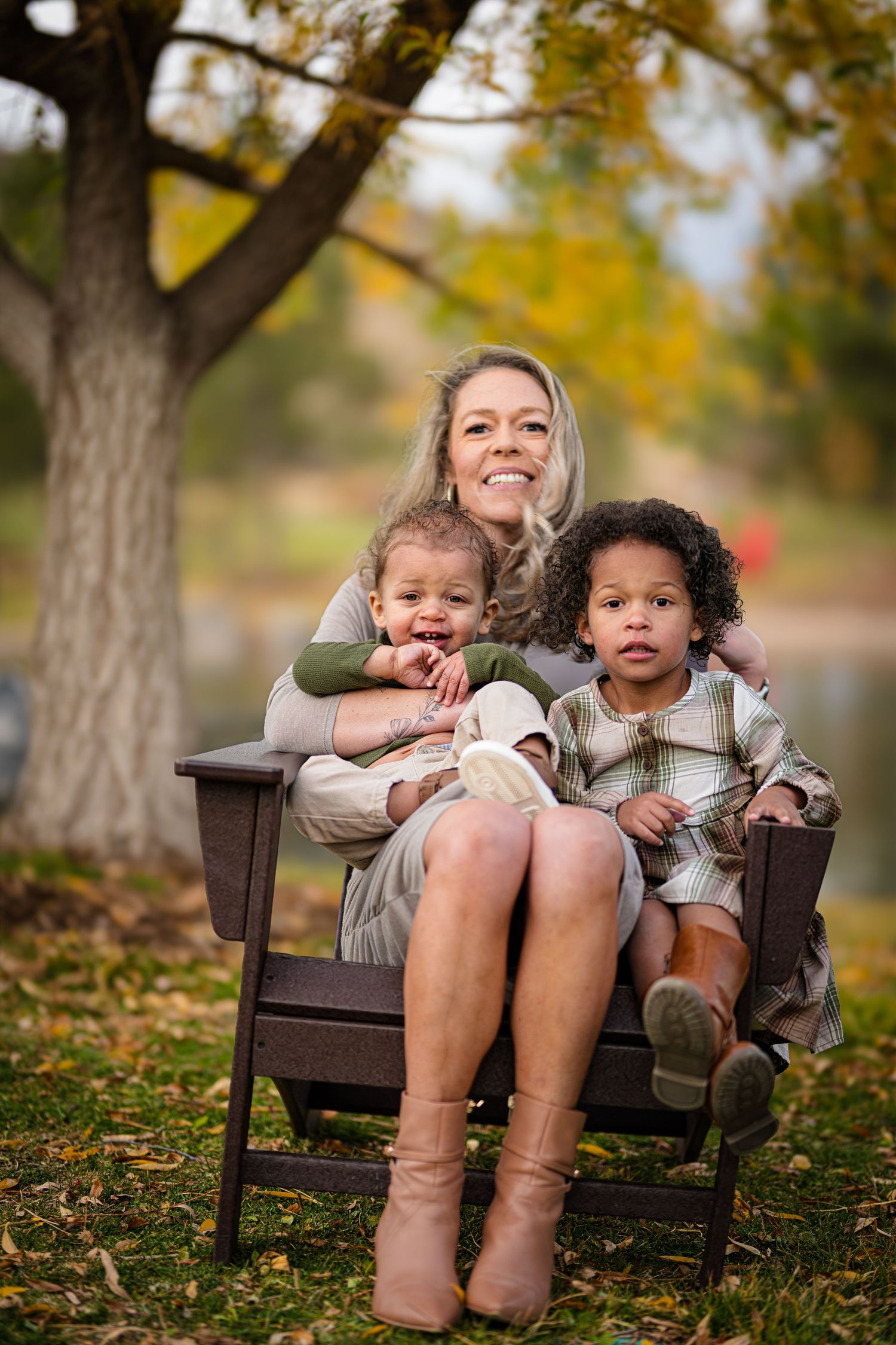 A blonde mother sits in a wooden outdoor chair with two young children on her lap in an autumn park.
