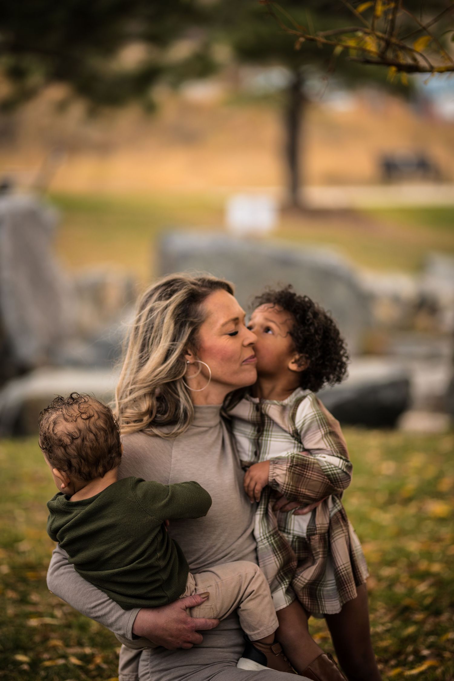 A loving mother receives a kiss from her toddler while holding her baby outdoors.