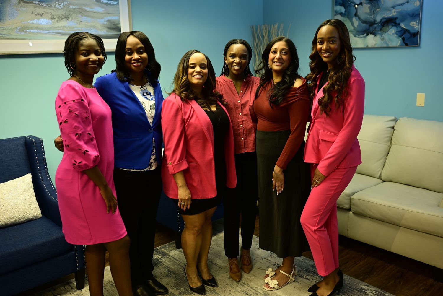 Six professional women smile together in a room with blue decor and a couch.