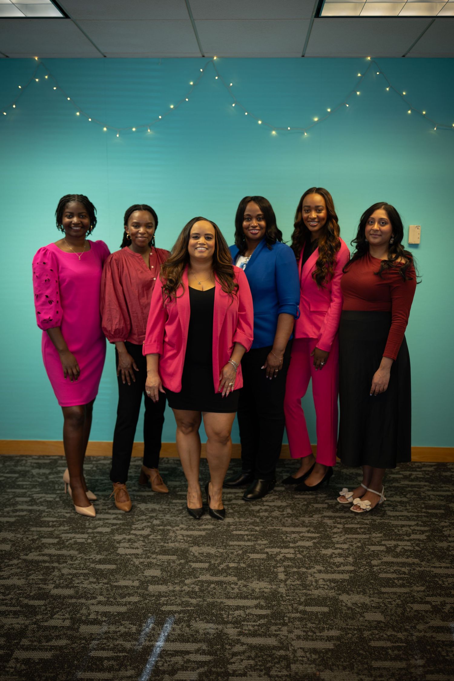 Six professional women in vibrant pink and blue outfits posing together against a teal wall with string lights.