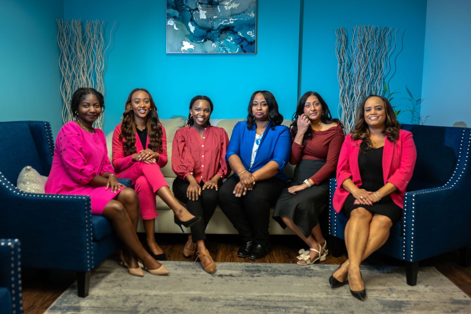 Six professional women in colorful outfits smiling and posing together on blue chairs in a teal room.