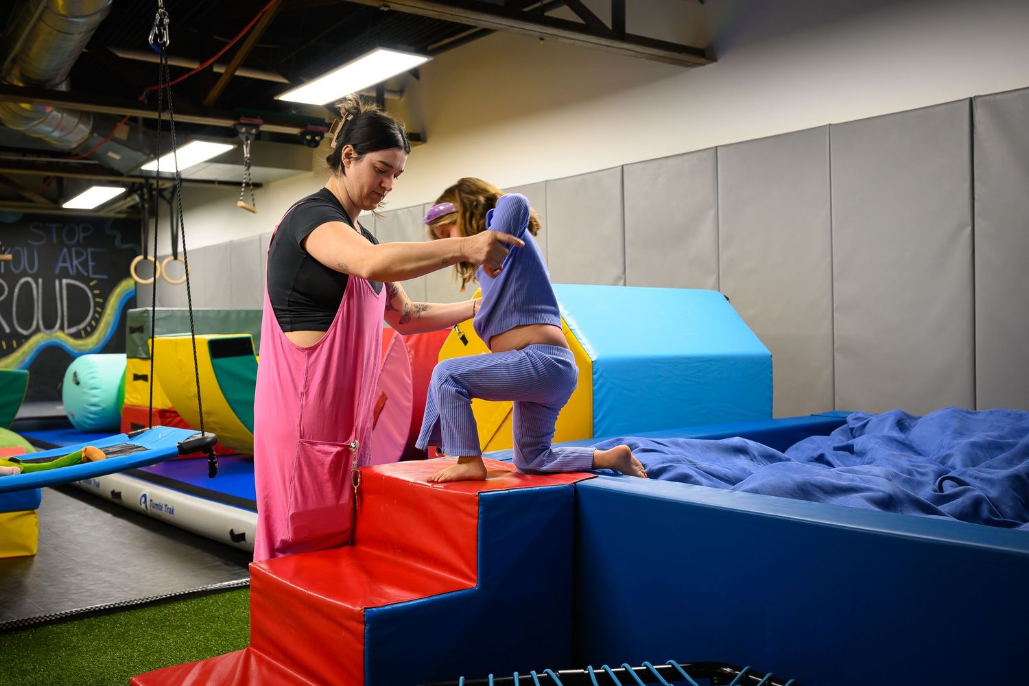 Female therapist in black top assists young child on colorful foam climbing equipment in sensory gym.