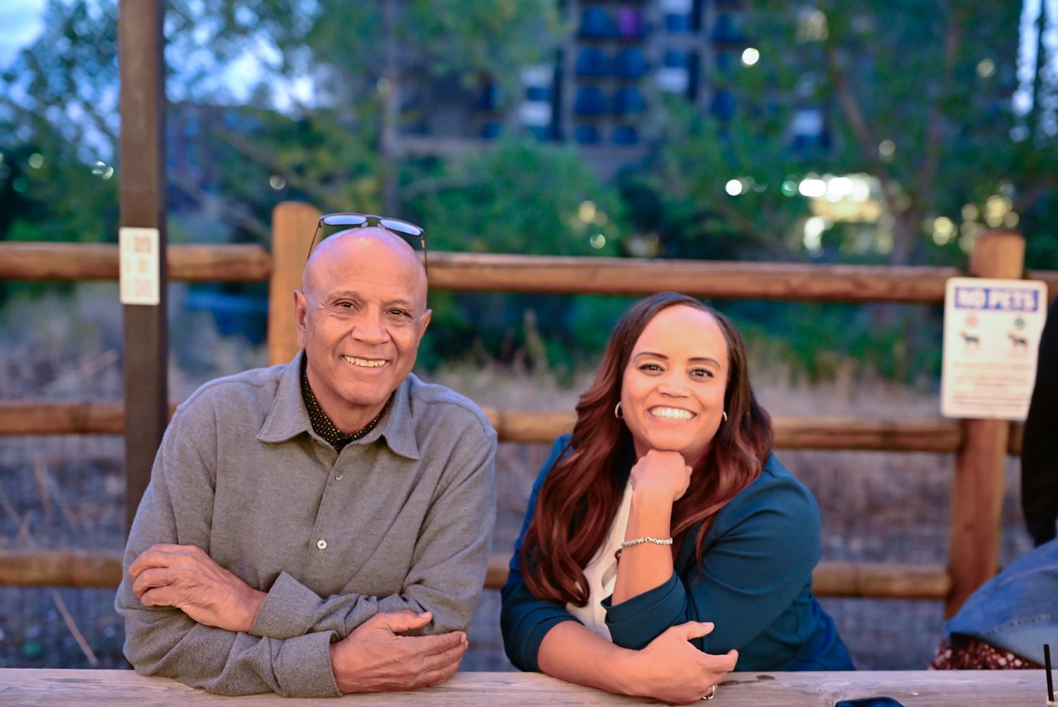 A man and woman smile together leaning on a wooden fence outdoors at dusk.