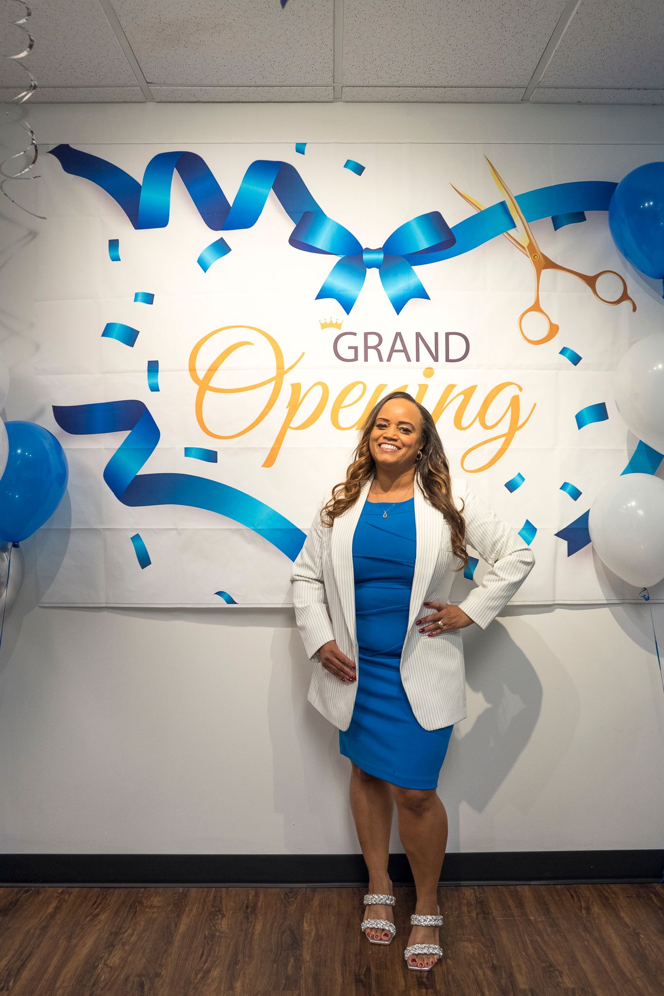 Smiling woman in blue dress and white blazer stands before a Grand Opening banner with balloons.