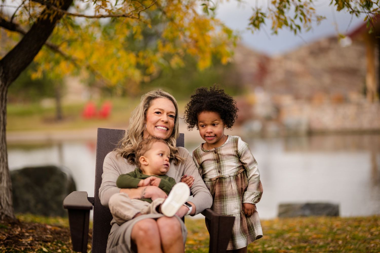 A smiling blonde mother sits outdoors with two young children in a colorful autumn park setting.