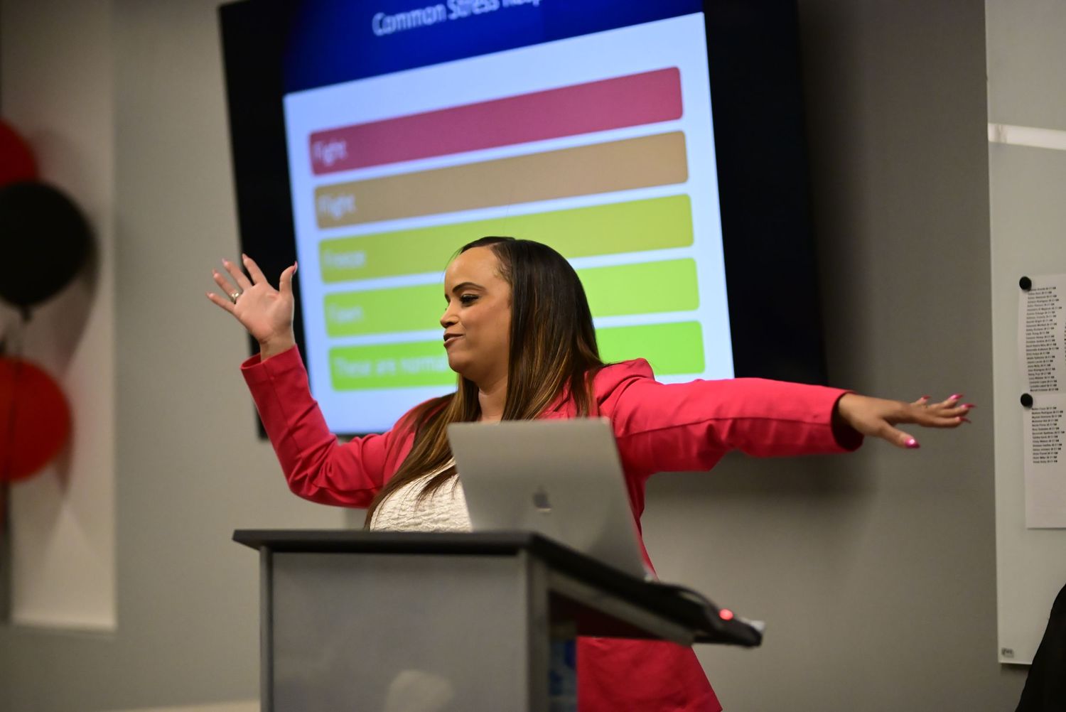 A woman in a red blazer presents with arms outstretched at a conference podium.