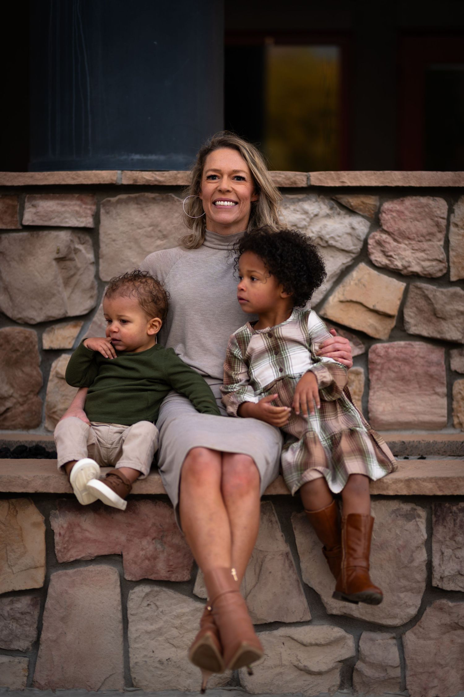 A smiling blonde woman sits on stone steps outdoors with two young mixed-race children in autumn attire.