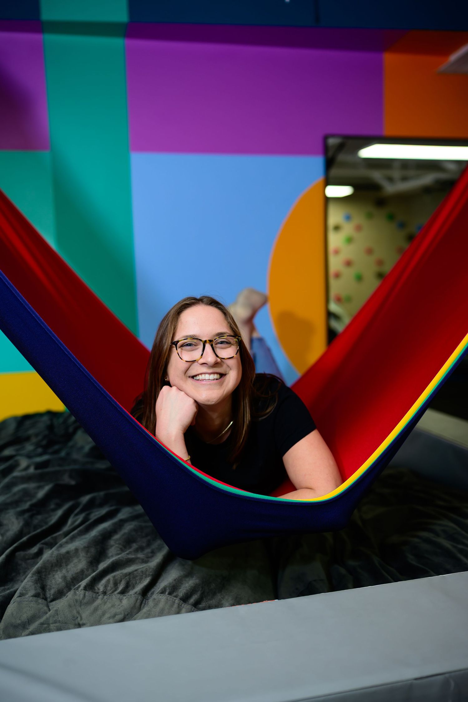 Smiling young woman with glasses relaxing in a colorful red, blue, and yellow hammock in vibrant room.