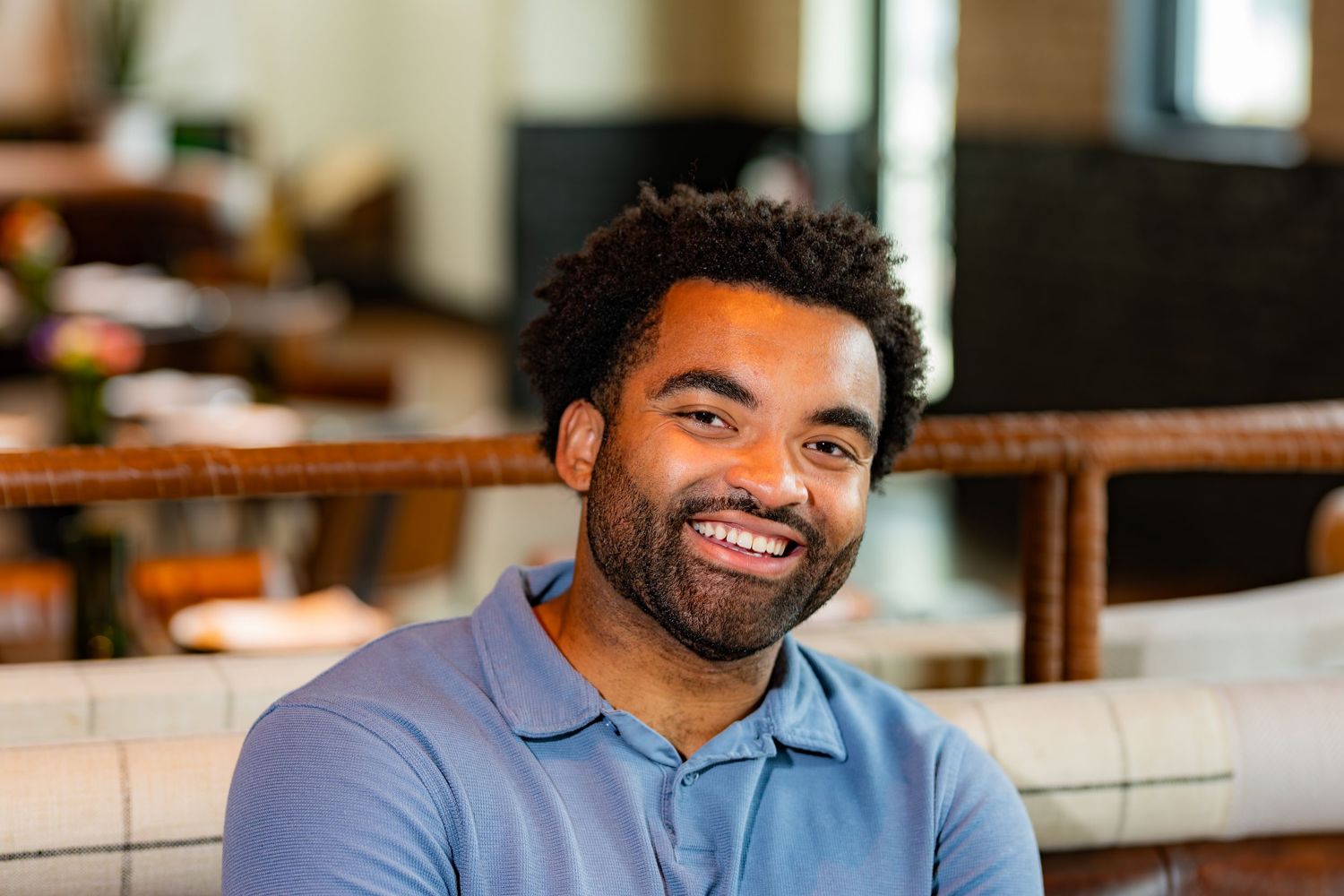 Smiling young Black man in blue polo shirt seated in casual restaurant setting.