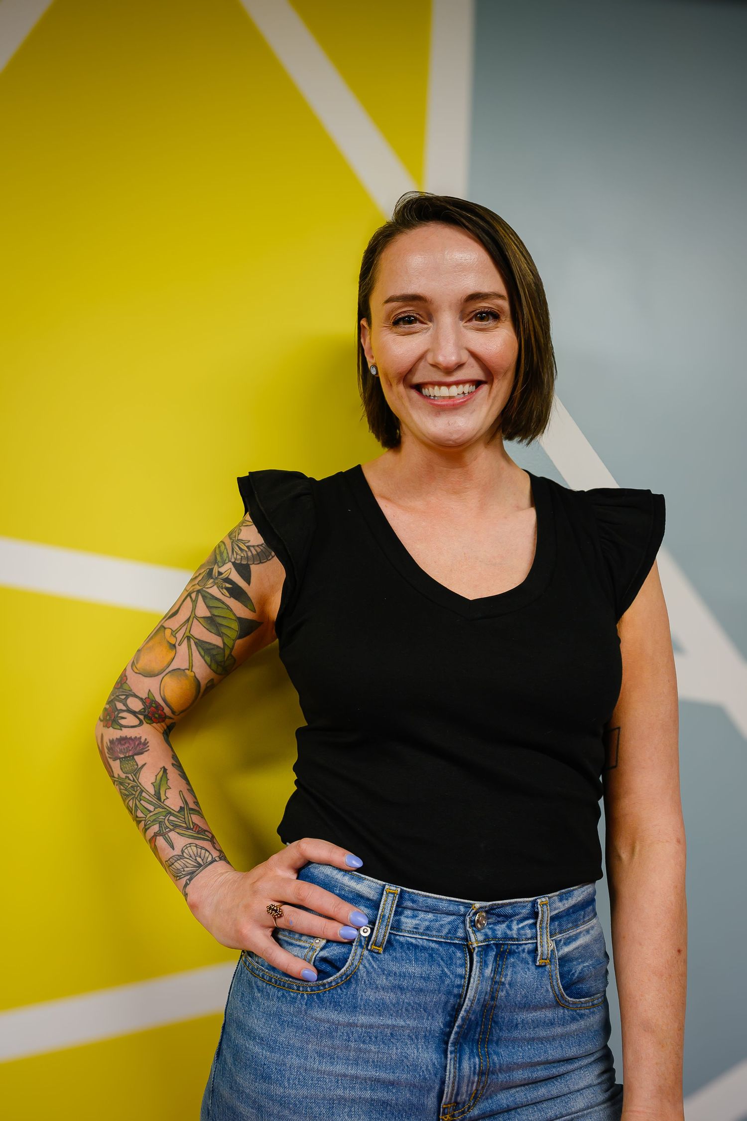 Smiling woman with floral sleeve tattoo in black top and jeans posing against yellow geometric wall.