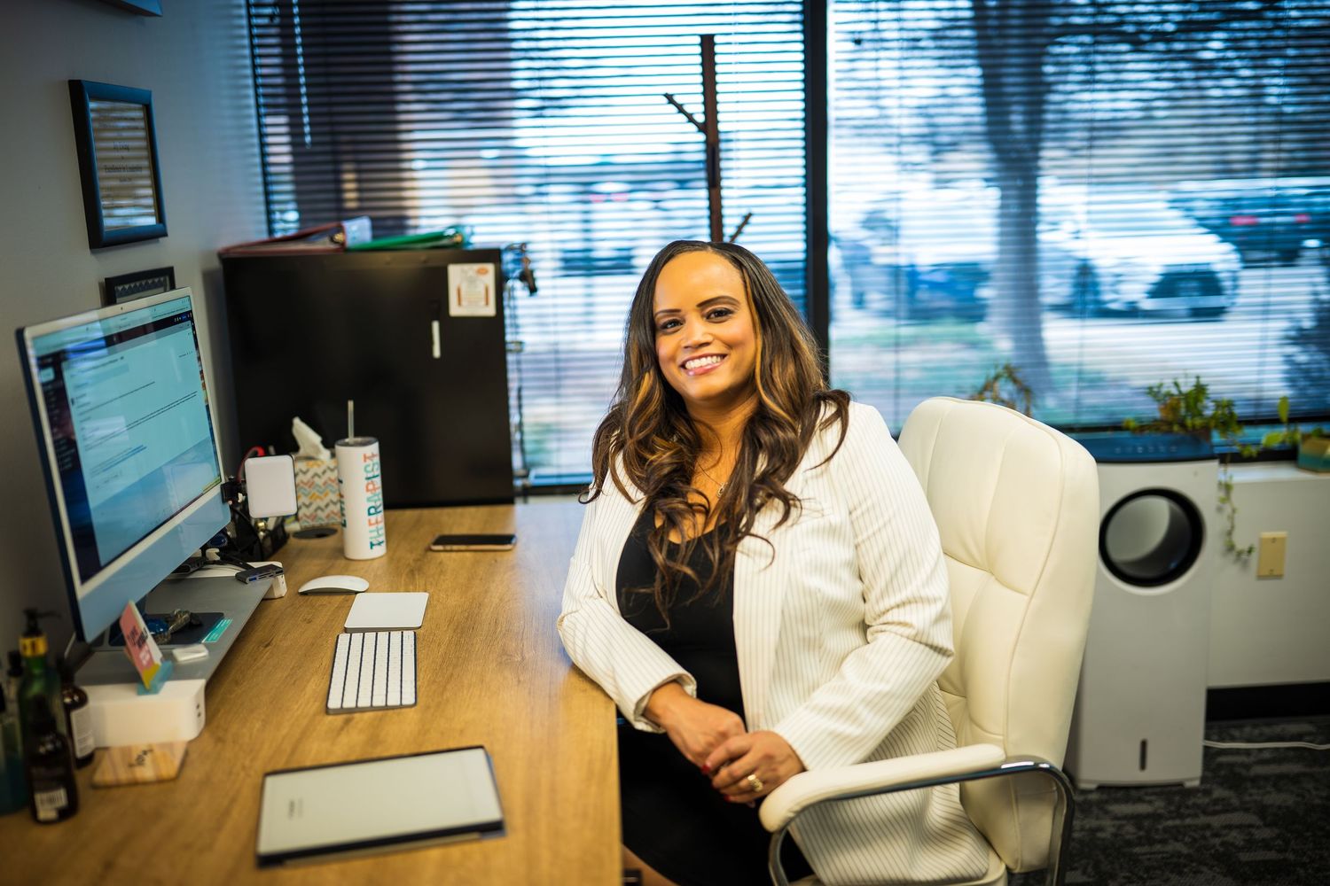 A professional woman in a white coat smiles while seated at a modern office desk with a computer.