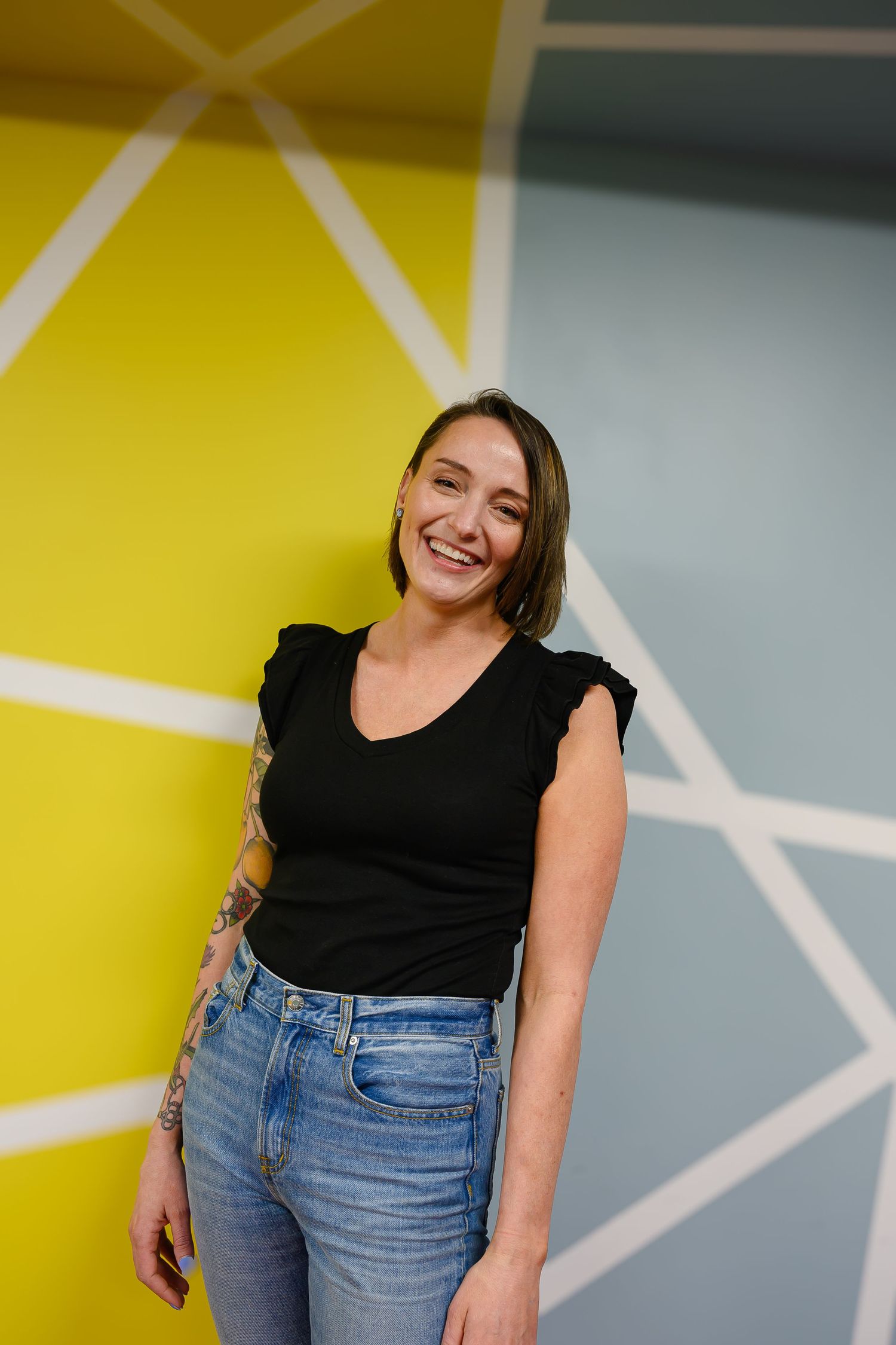 A smiling woman in a black top and jeans poses casually against a colorful geometric yellow and gray wall.