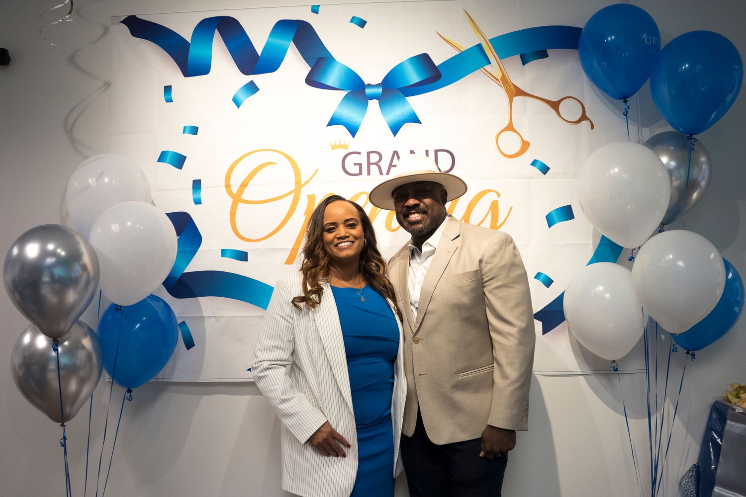 A couple poses together at a grand opening celebration with blue and white balloons and confetti decorations.