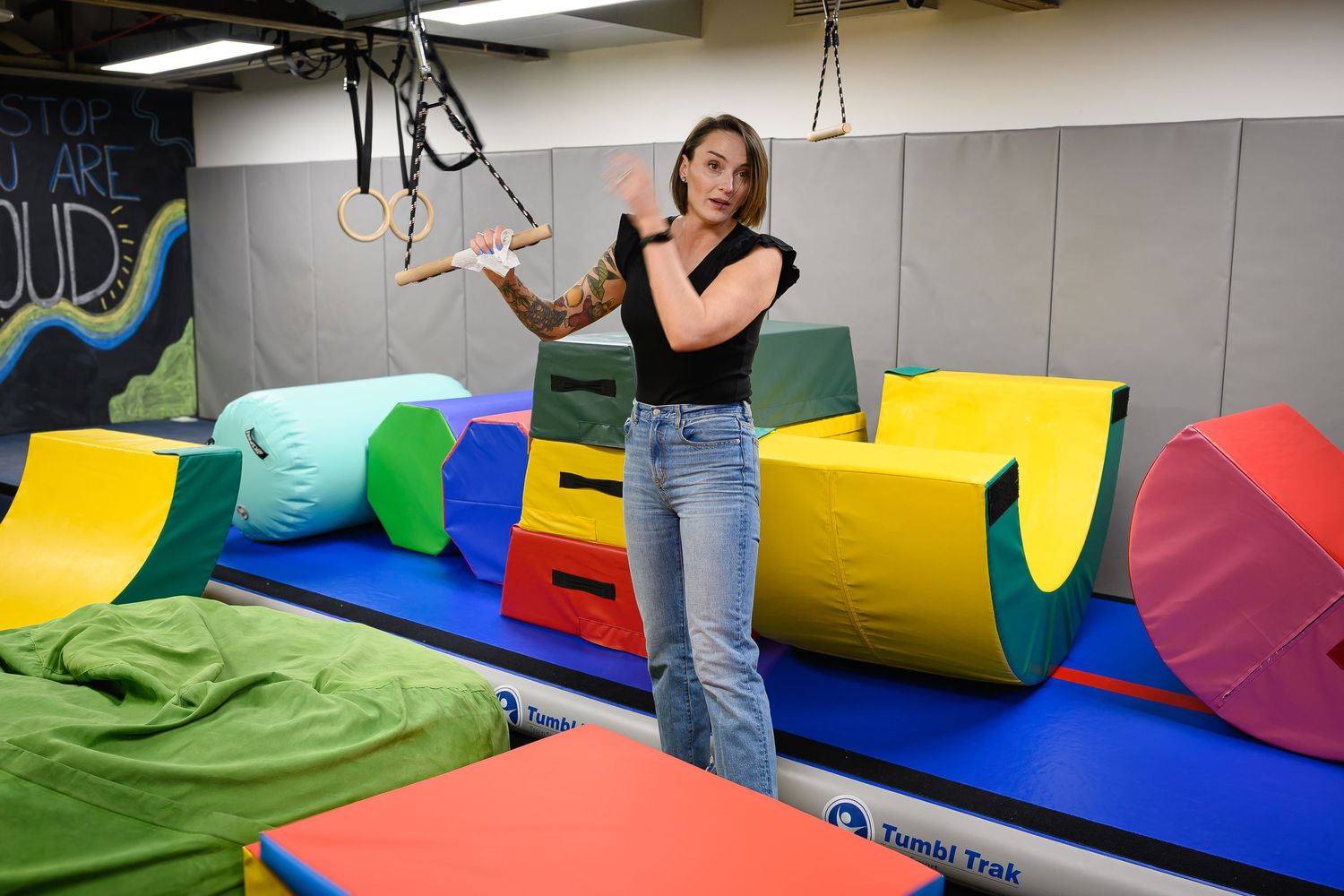 Woman with tattoos standing among colorful foam play equipment in children's therapy gym.