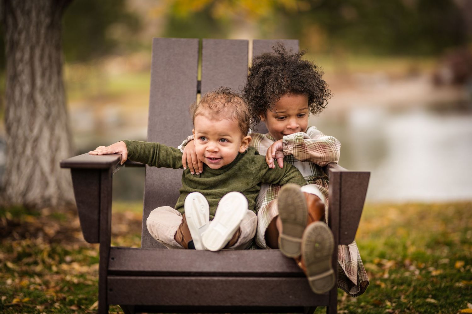 Two young siblings sit together smiling in a wooden Adirondack chair outdoors.