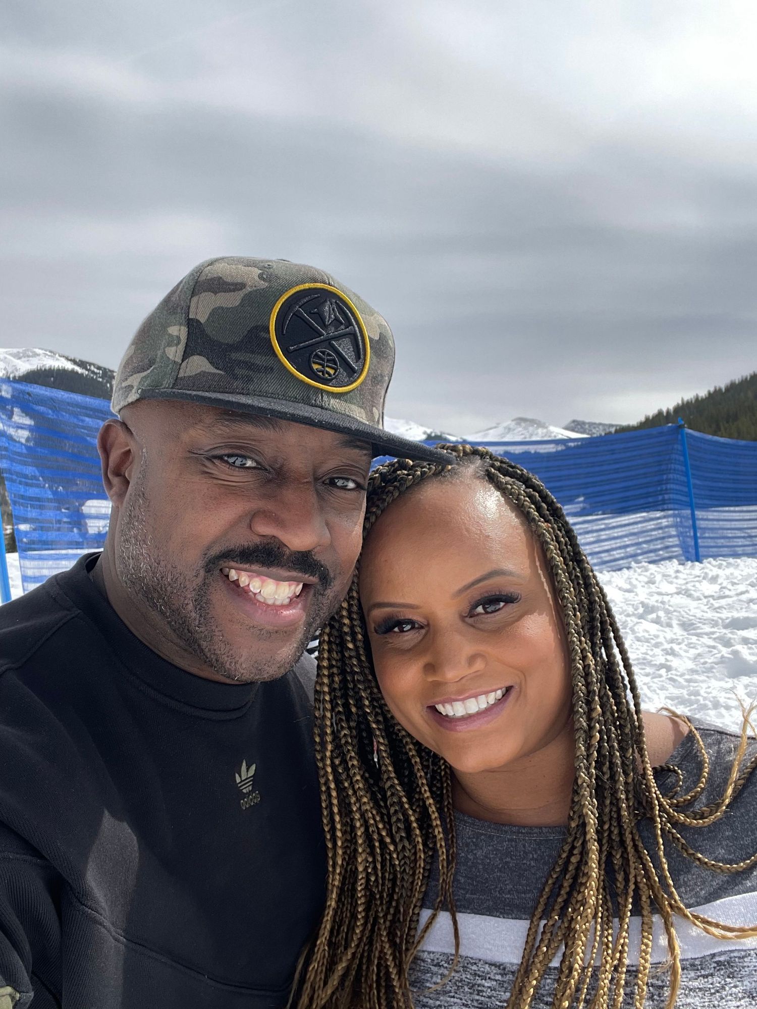 A smiling couple poses together outdoors in a snowy mountain setting wearing casual winter attire.