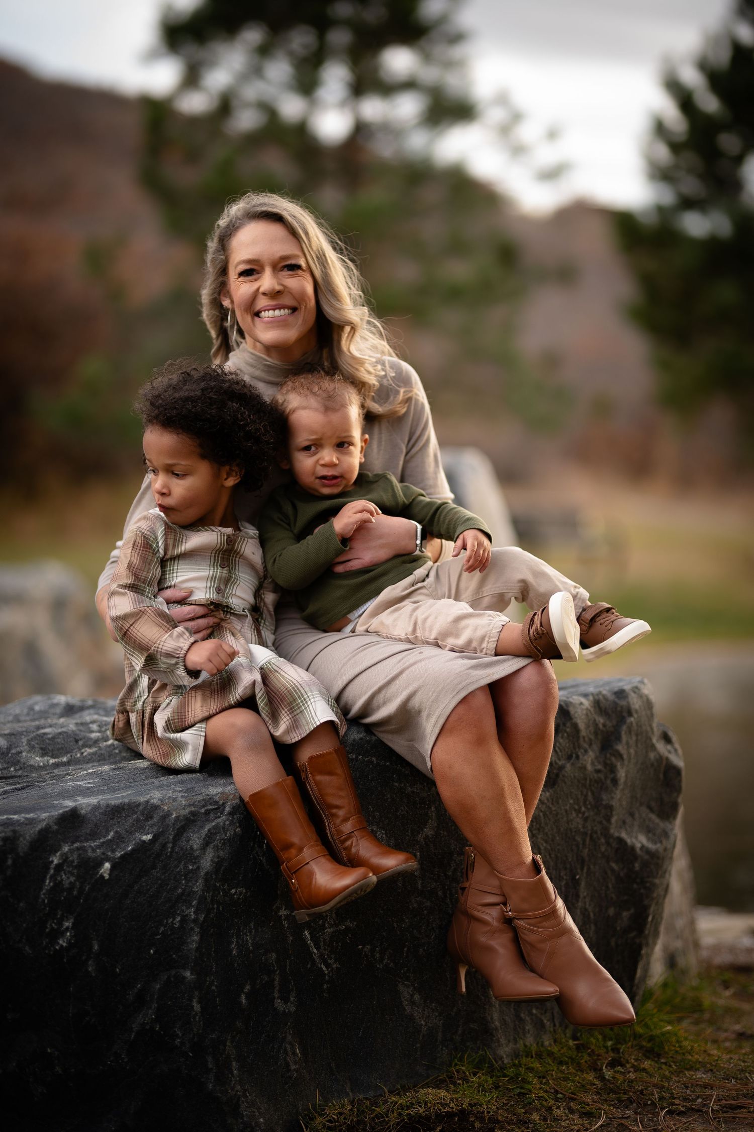 A happy mother sits on a rock with her two young children in an outdoor setting.