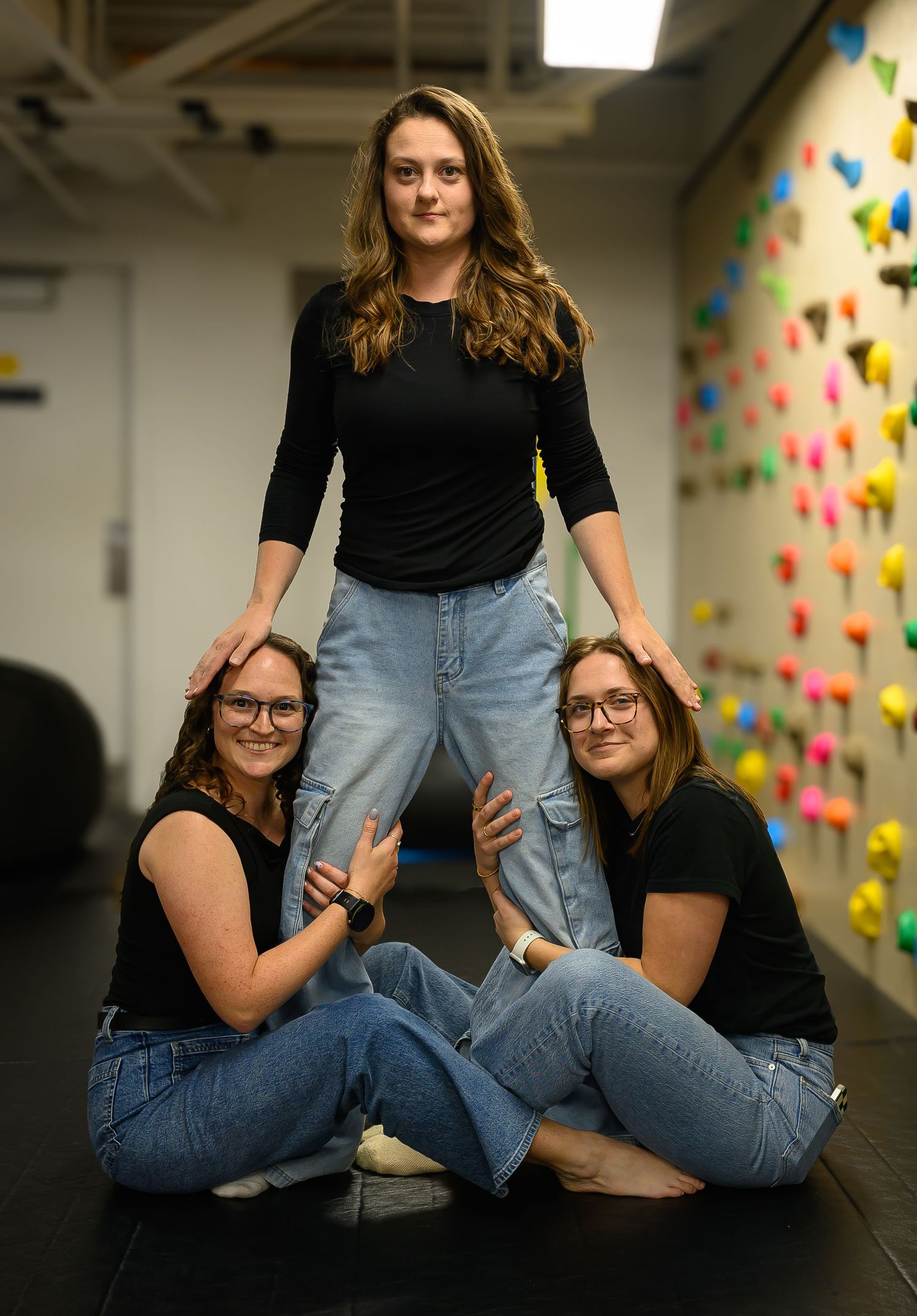 Three women posing playfully in front of colorful climbing wall in therapy gym.
